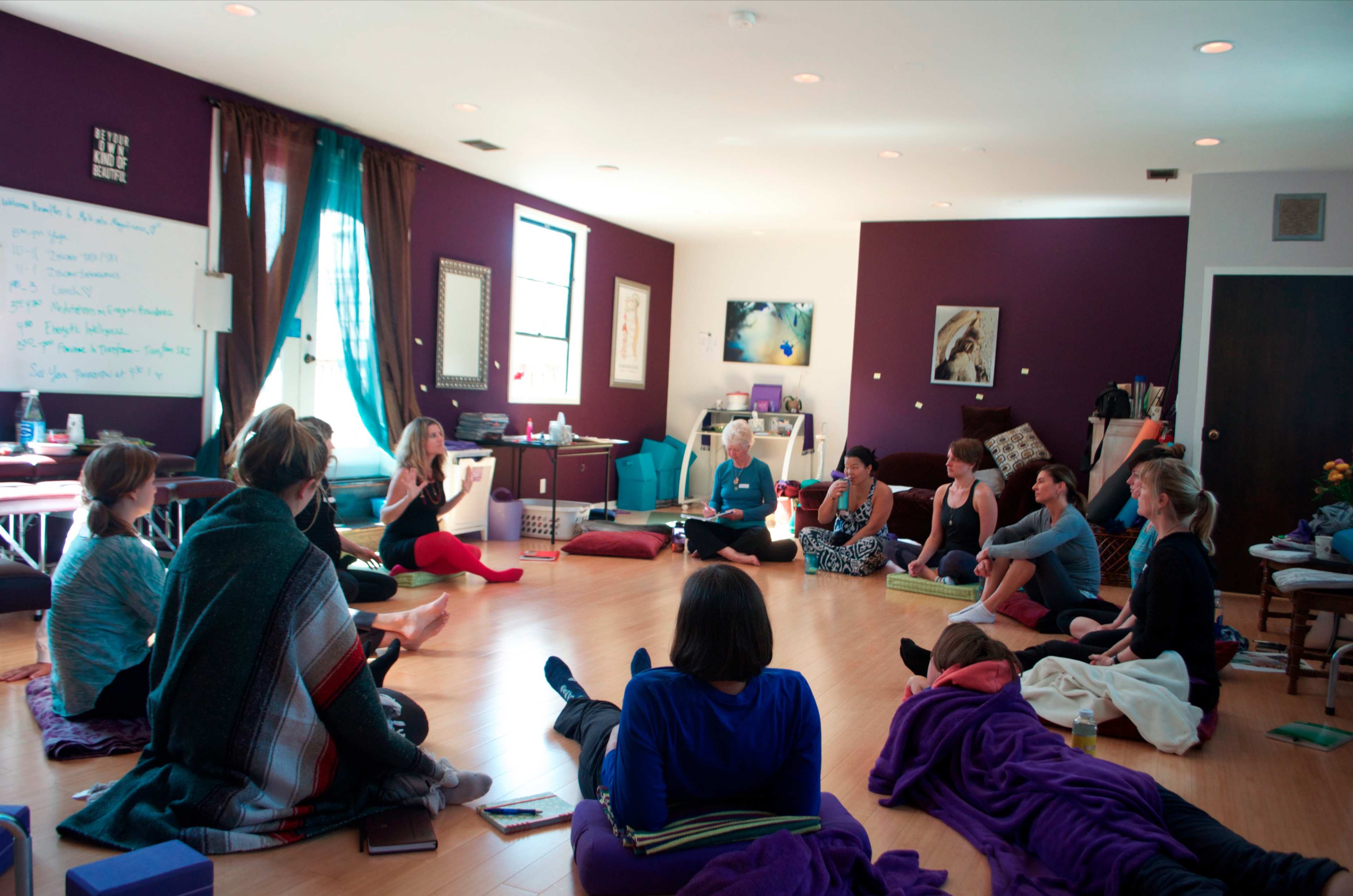 A group of people sit in a circle on the floor of a room with purple walls, engaged in a discussion.