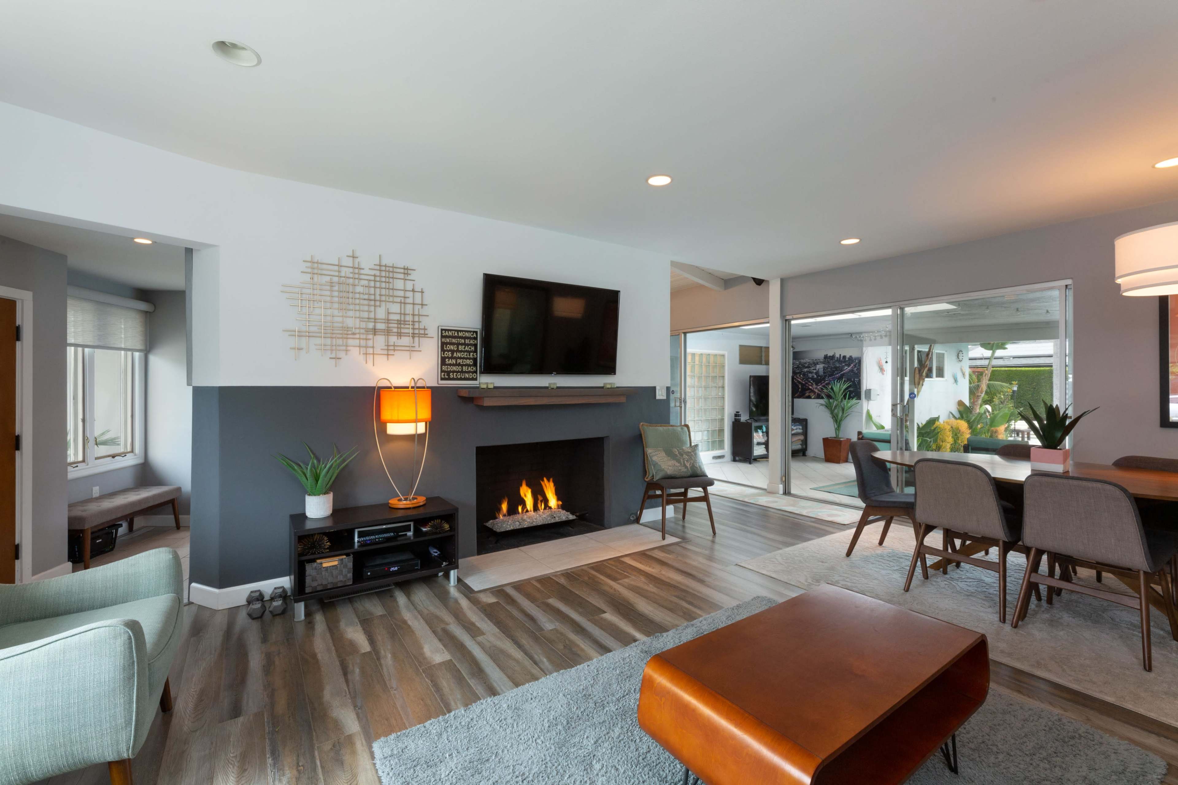 A modern living room featuring a wall-mounted TV above a fireplace, with a dining area in the background and large windows that extend into an outdoor space.