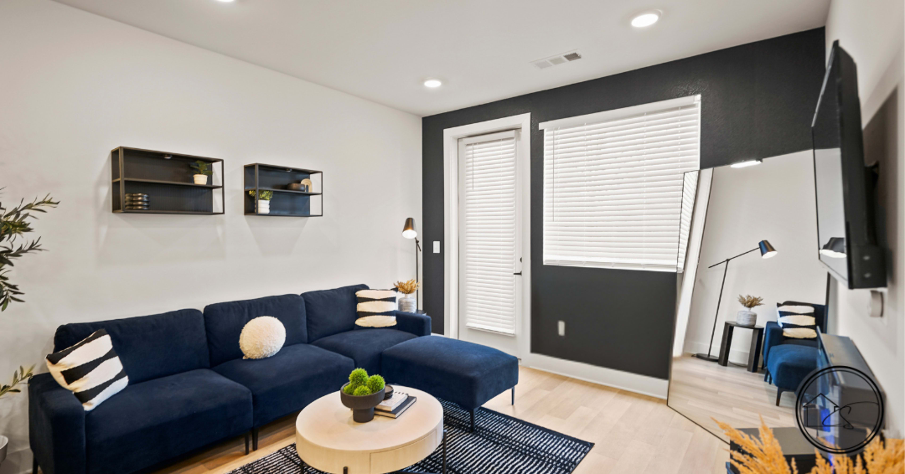 The image shows a modern living room with a blue sectional sofa, a round coffee table, and a wall-mounted television, featuring large windows with blinds and decorative shelves.