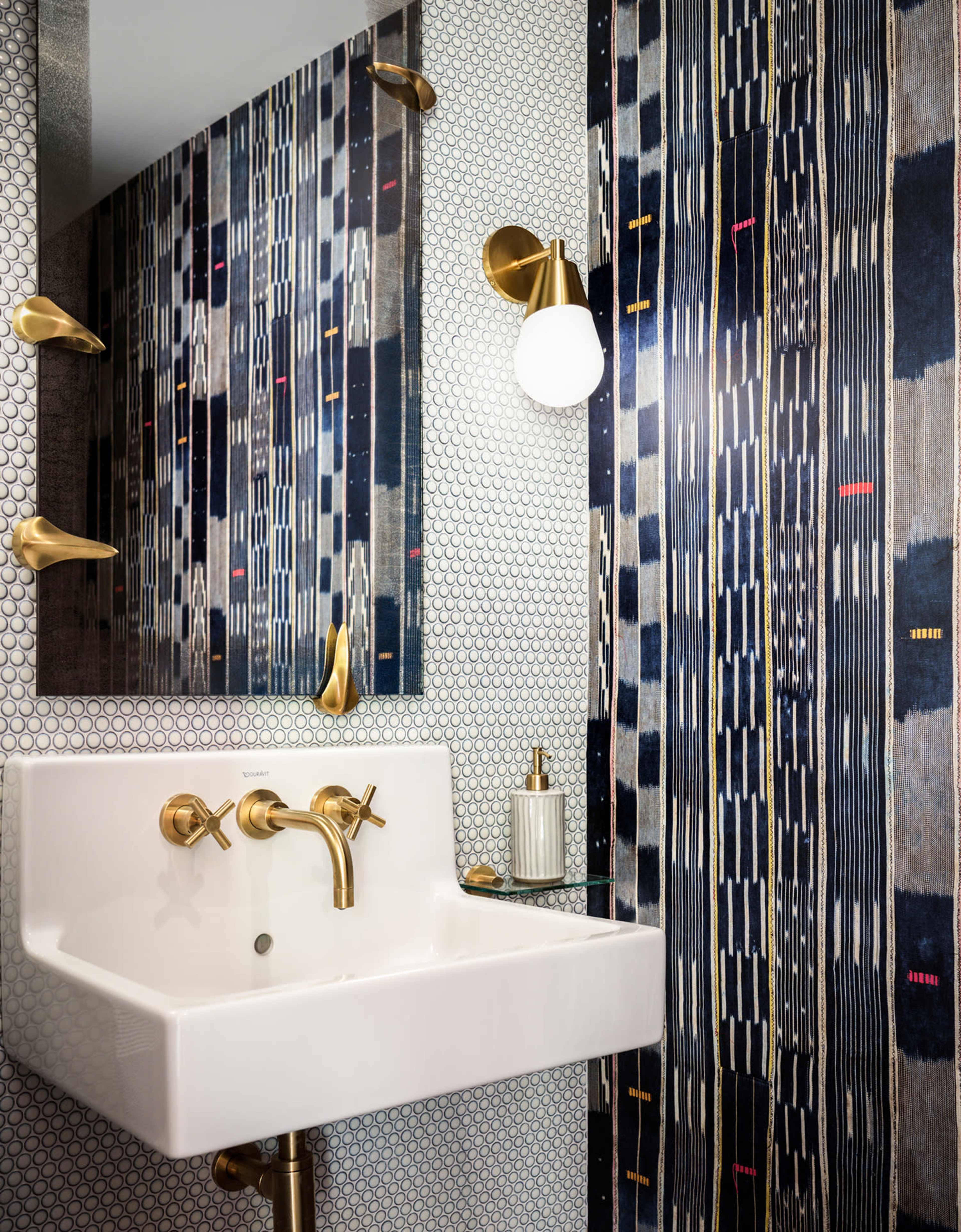 The image shows a modern bathroom with a white sink, brass fixtures, and patterned wallpaper featuring vertical lines in blue and black hues.