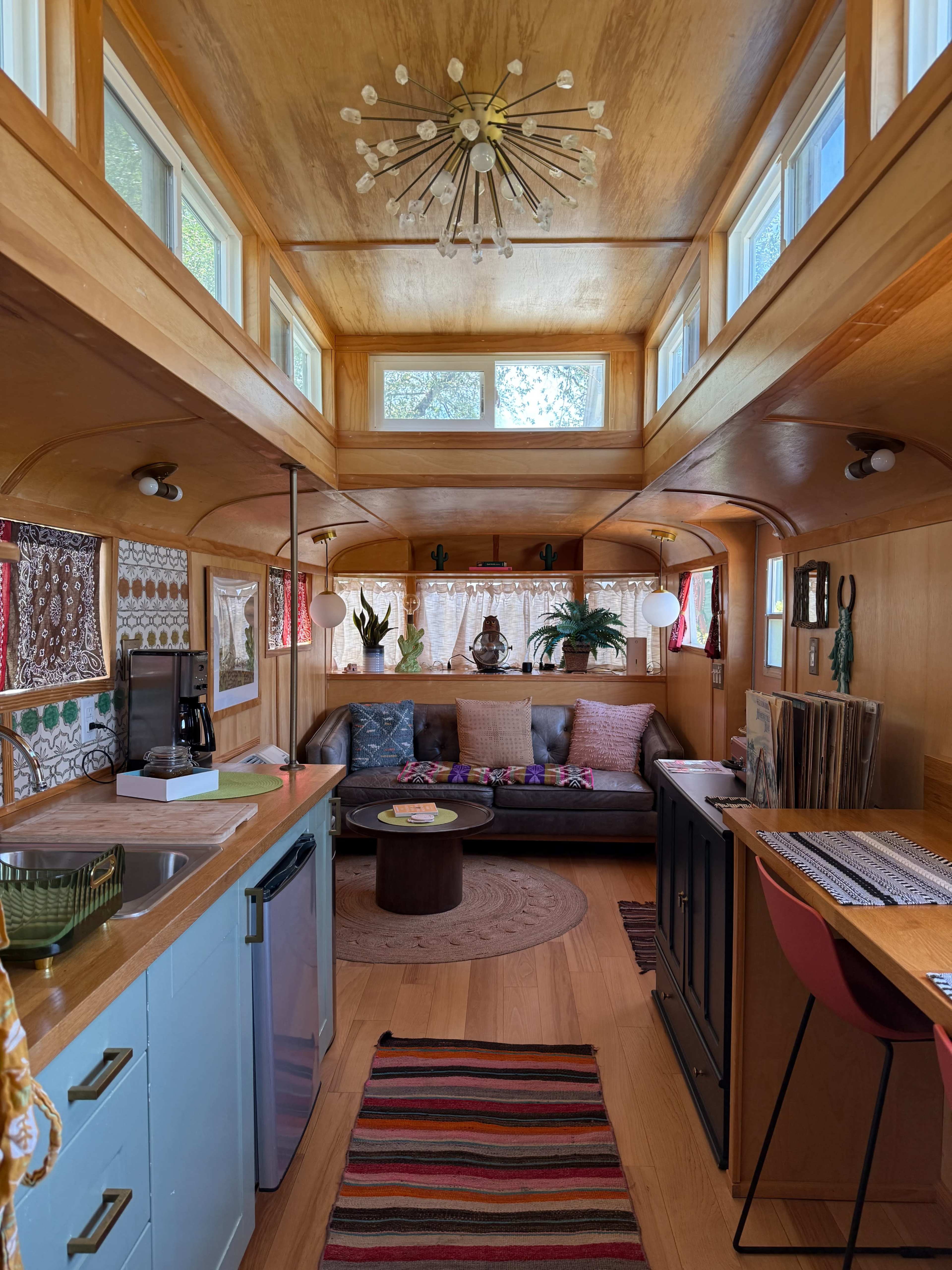 The interior of a renovated camper features wood-paneled walls, a cozy seating area with a small table, a kitchenette, and large windows allowing natural light to brighten the space.