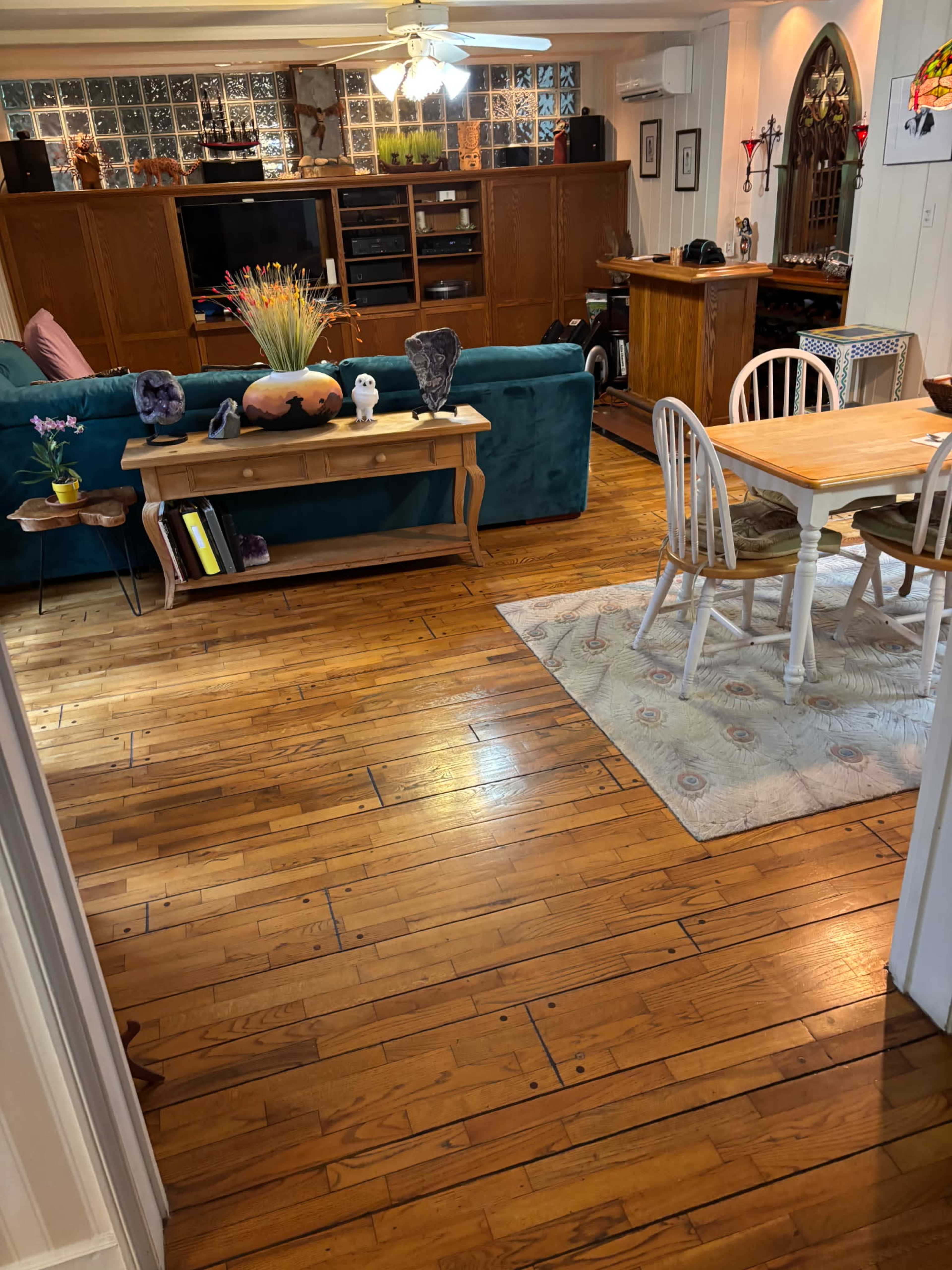 The image shows a living and dining area with wooden floors, a green couch, a wooden dining table, and various decorative items on the furniture.