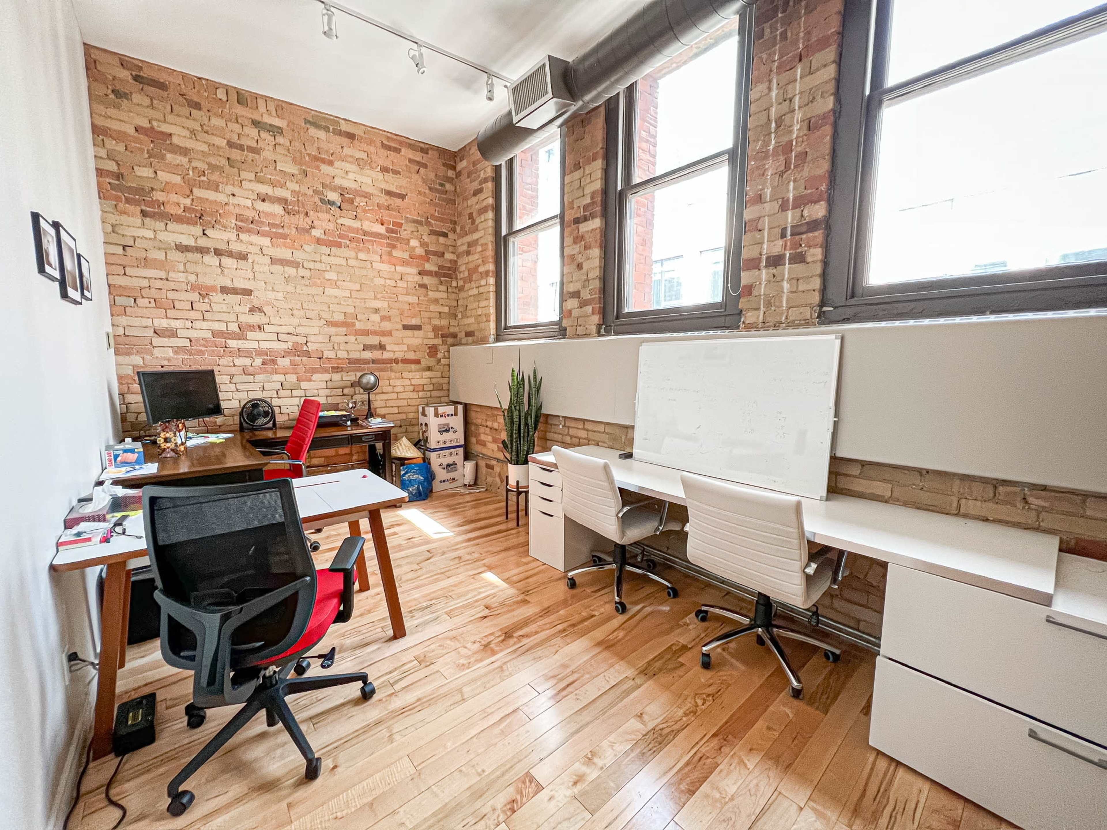 A modern office space with exposed brick walls, three desks, and large windows providing natural light.