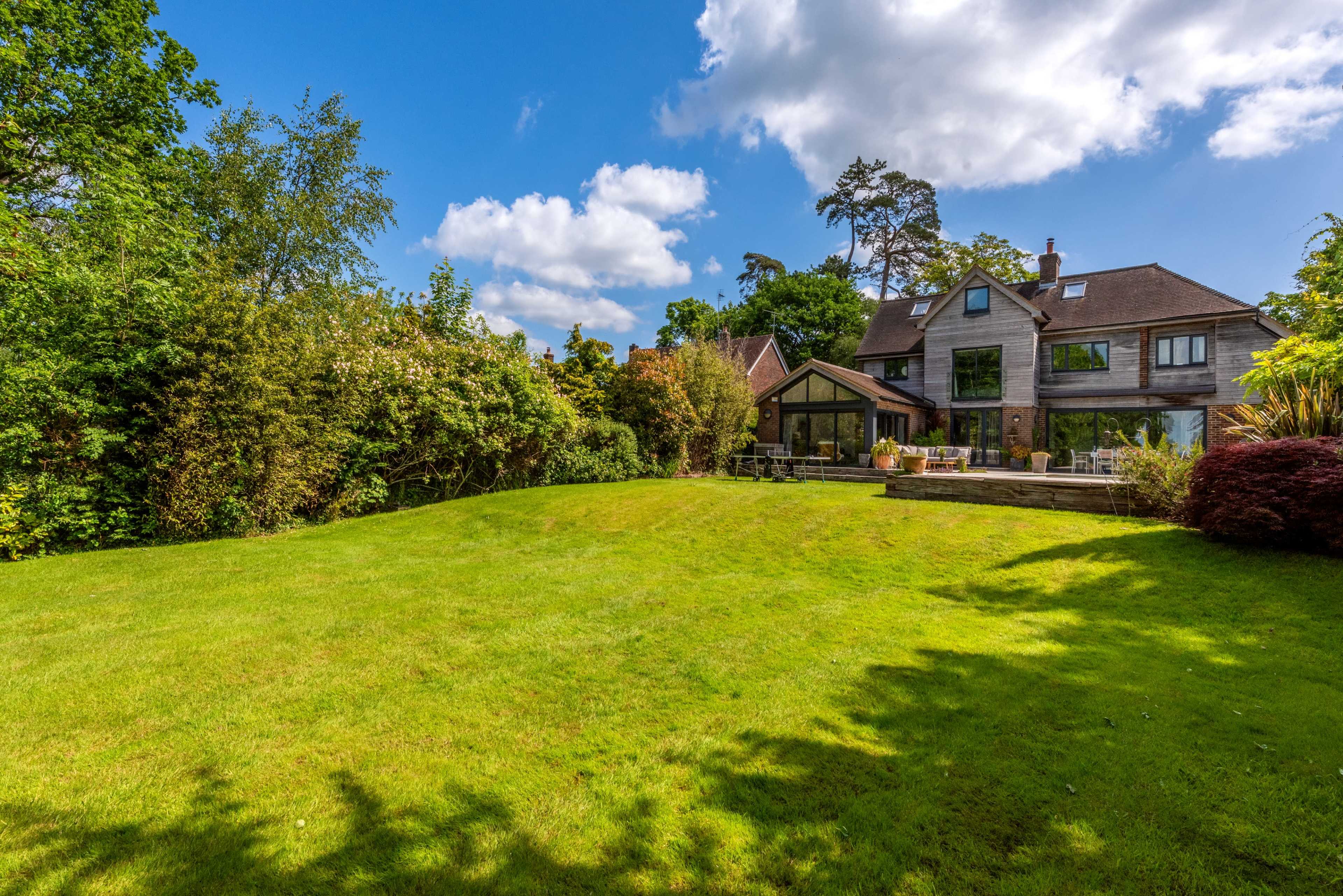A well-maintained lawn in front of a large two-story house surrounded by trees and shrubs under a clear blue sky with scattered clouds.