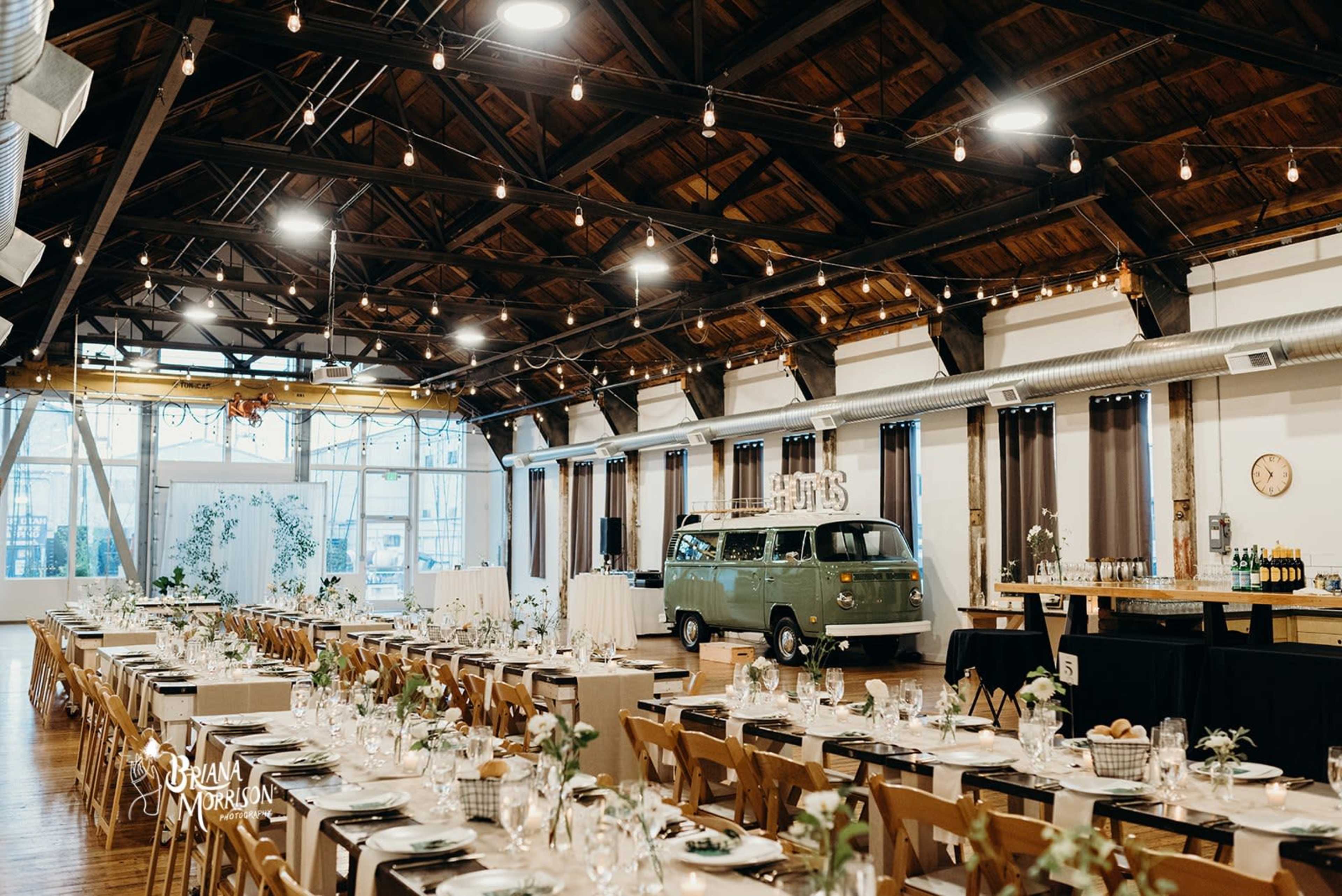 A spacious event venue features long reception tables set for a banquet, with a vintage green van parked at one end and decorative string lights overhead.