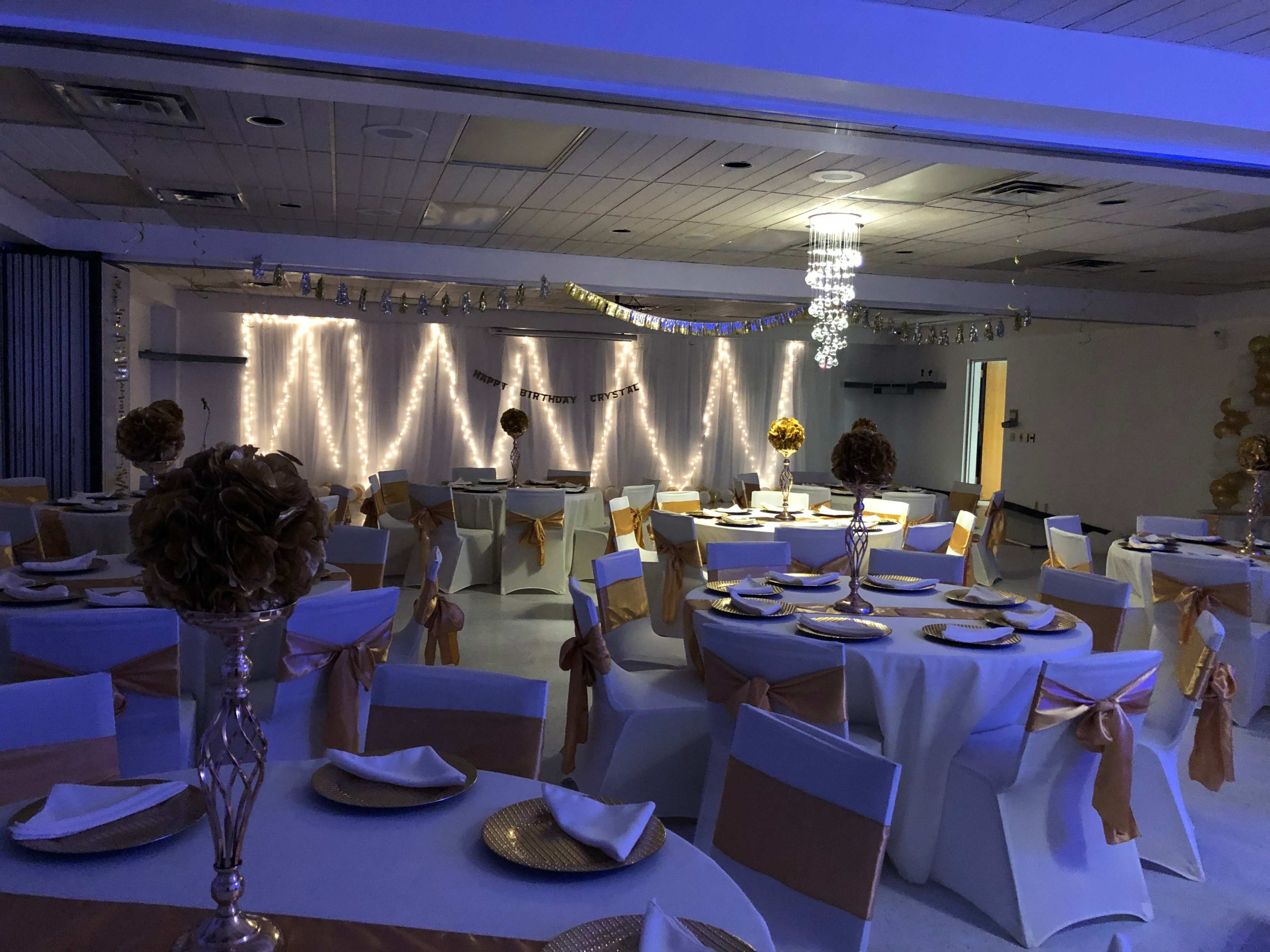 The interior of a banquet hall is set with round tables covered in white tablecloths and adorned with gold accents, illuminated by blue lighting and decorative string lights.