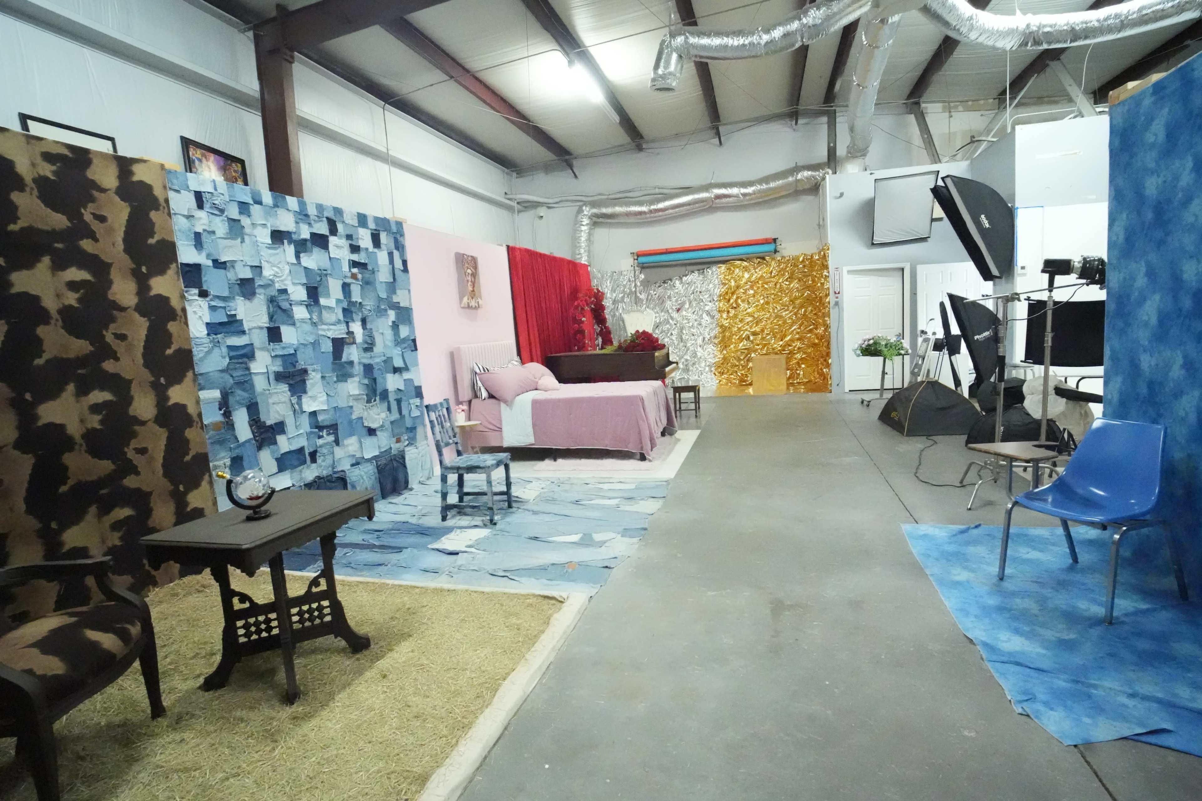 The image shows a spacious studio with various themed backdrops, including textured walls in blue and gold, a pink wall with bedding, and a brown patterned area, along with furniture scattered throughout.