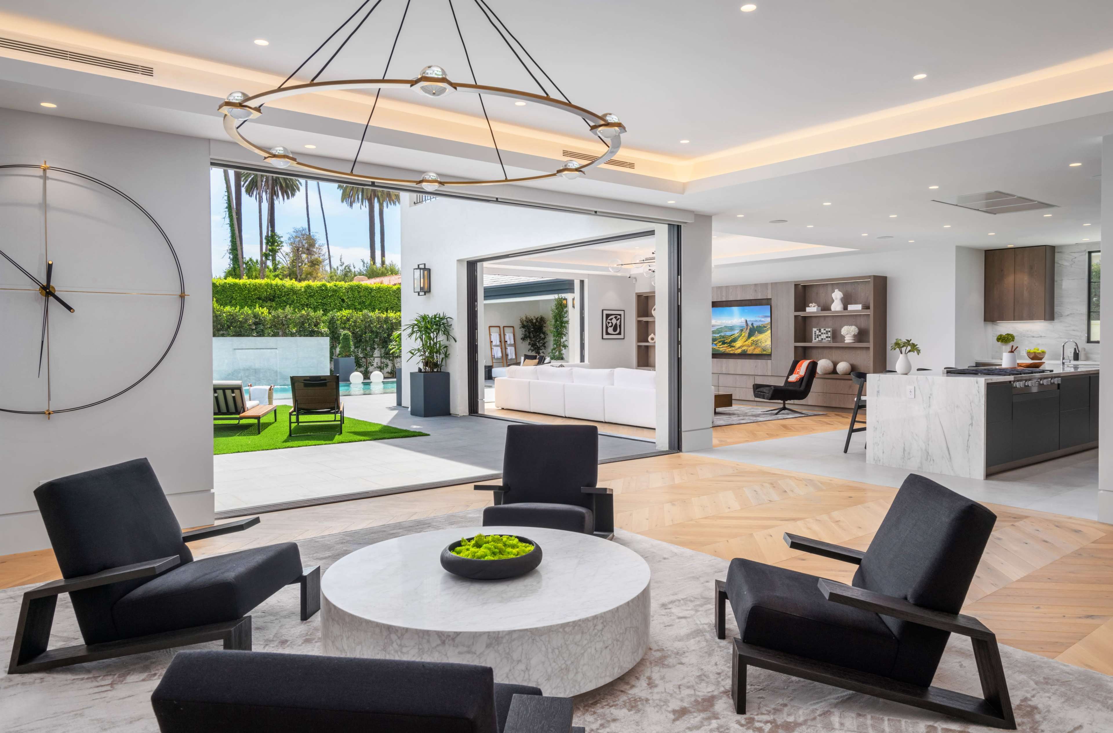 The image shows a modern living space featuring a circular coffee table surrounded by black lounge chairs, with an open view to an outdoor area and a kitchen in the background.