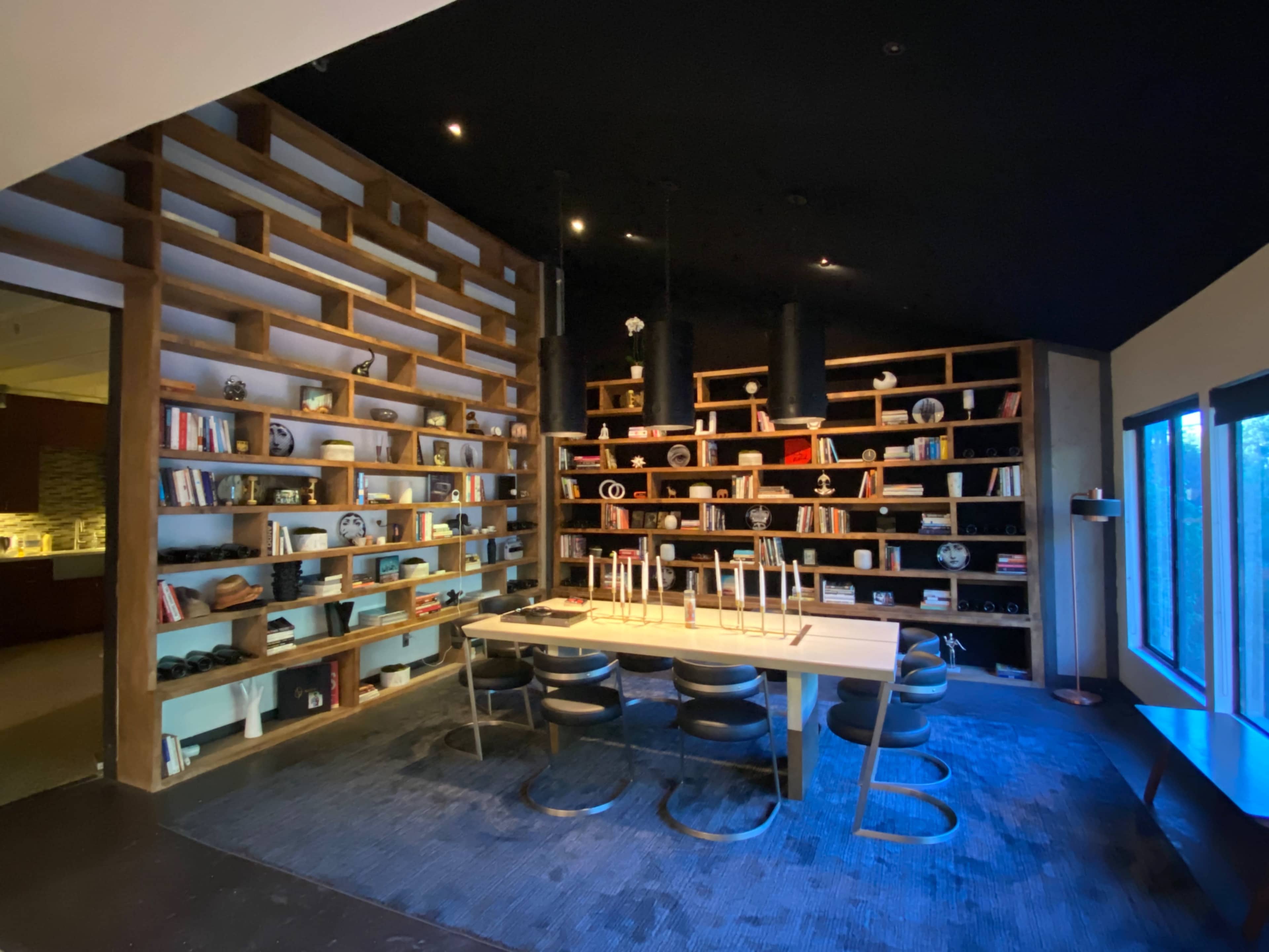 A modern dining room features a large wooden table surrounded by black chairs, with a wall of shelves displaying decorative objects and books.
