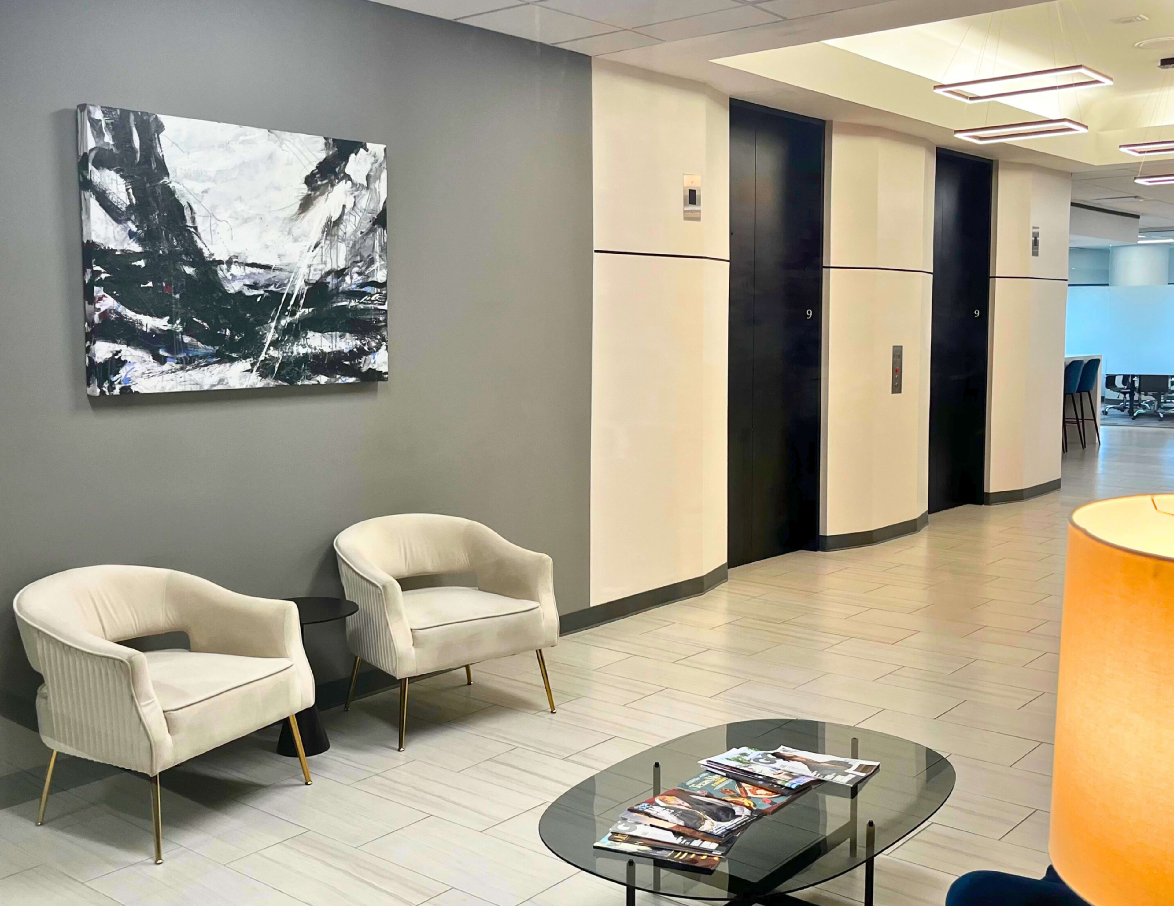 The scene depicts a modern lobby area featuring two white armchairs, a glass coffee table with magazines, and elevator doors along a sleek, light-colored corridor.
