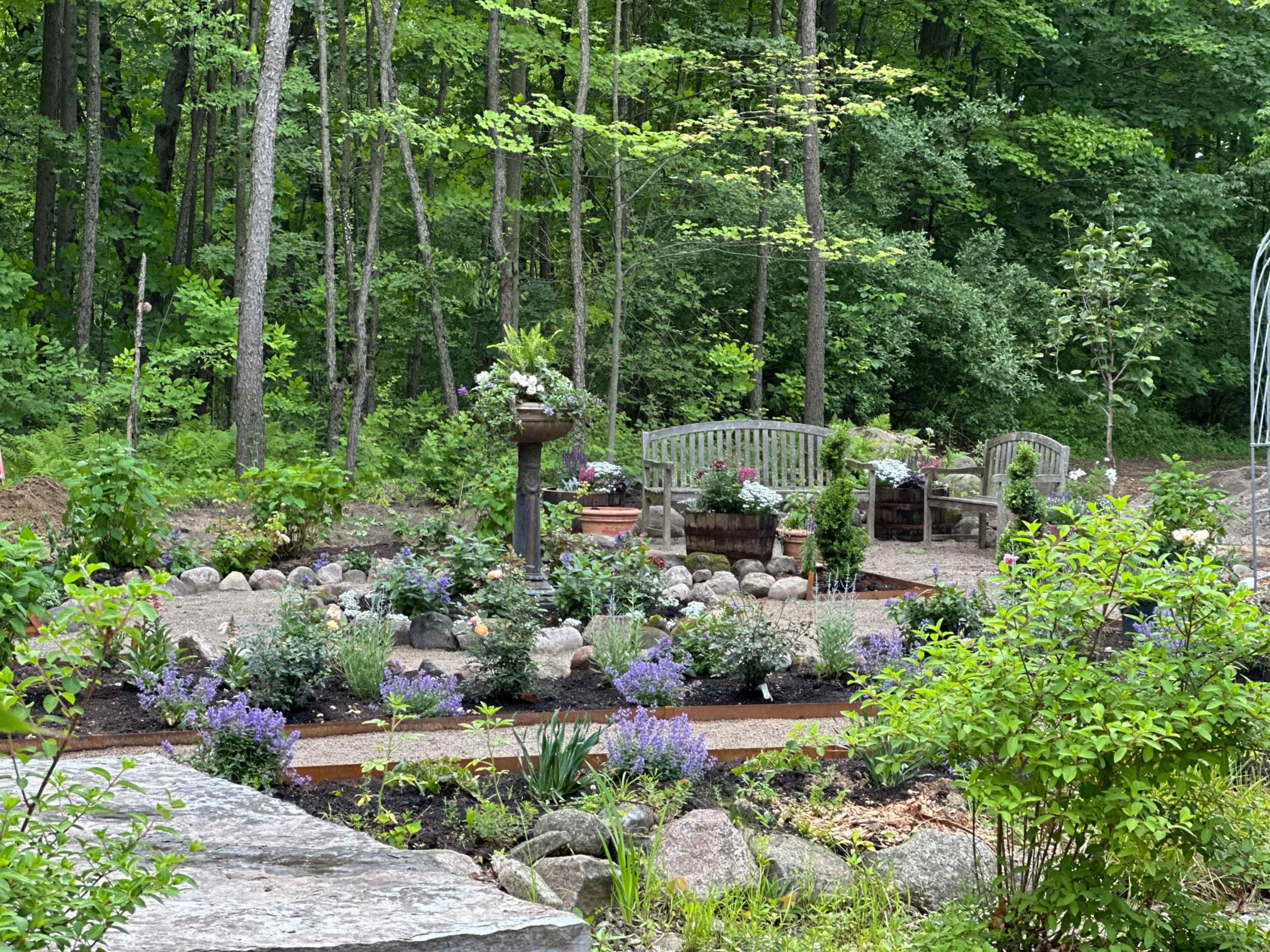 The scene features a landscaped garden with two wooden benches surrounded by flowerbeds and trees in the background.