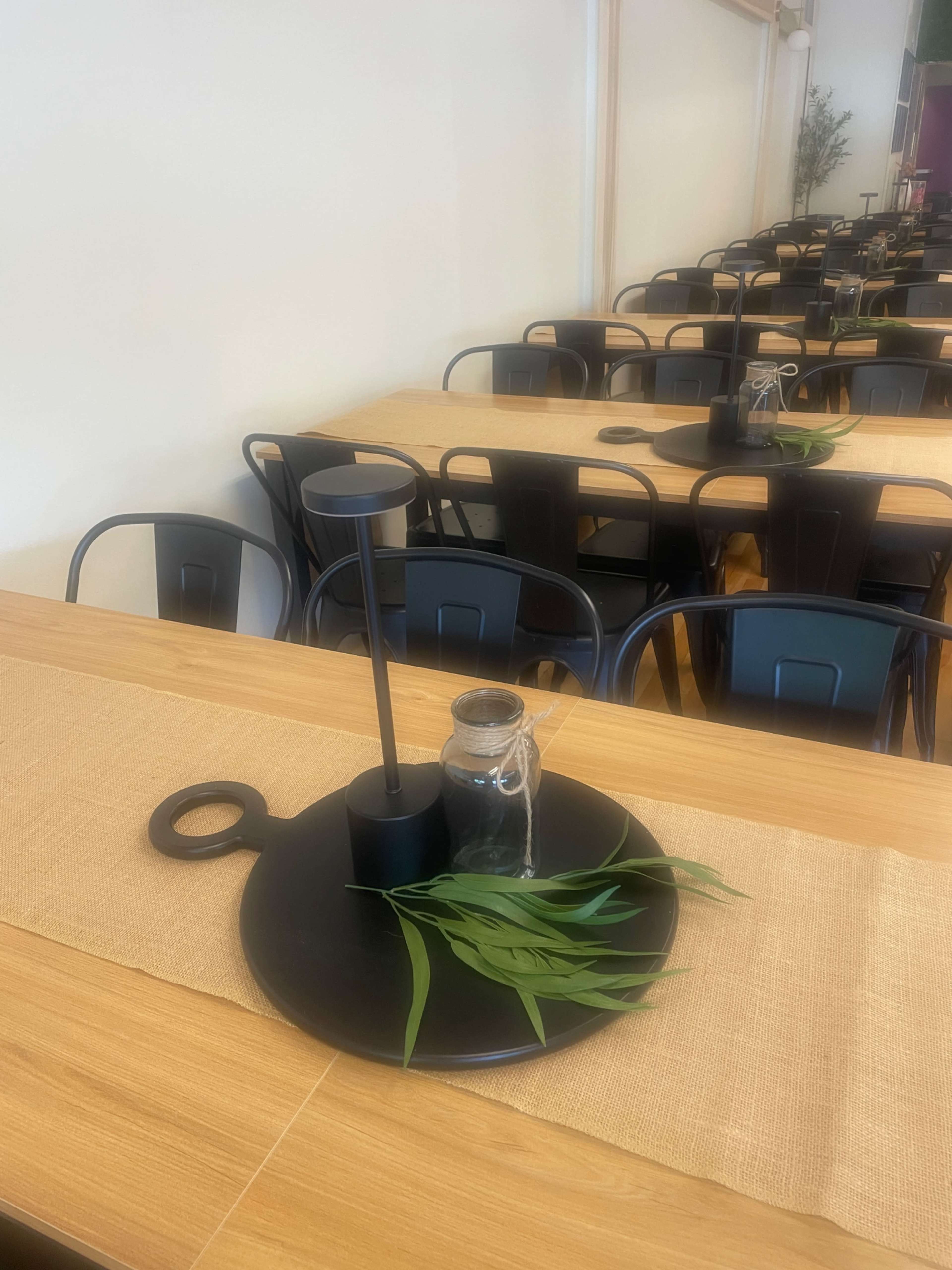 A table setting features a black tray with a candle holder, a small glass jar, and green leaves, arranged on a linen runner.