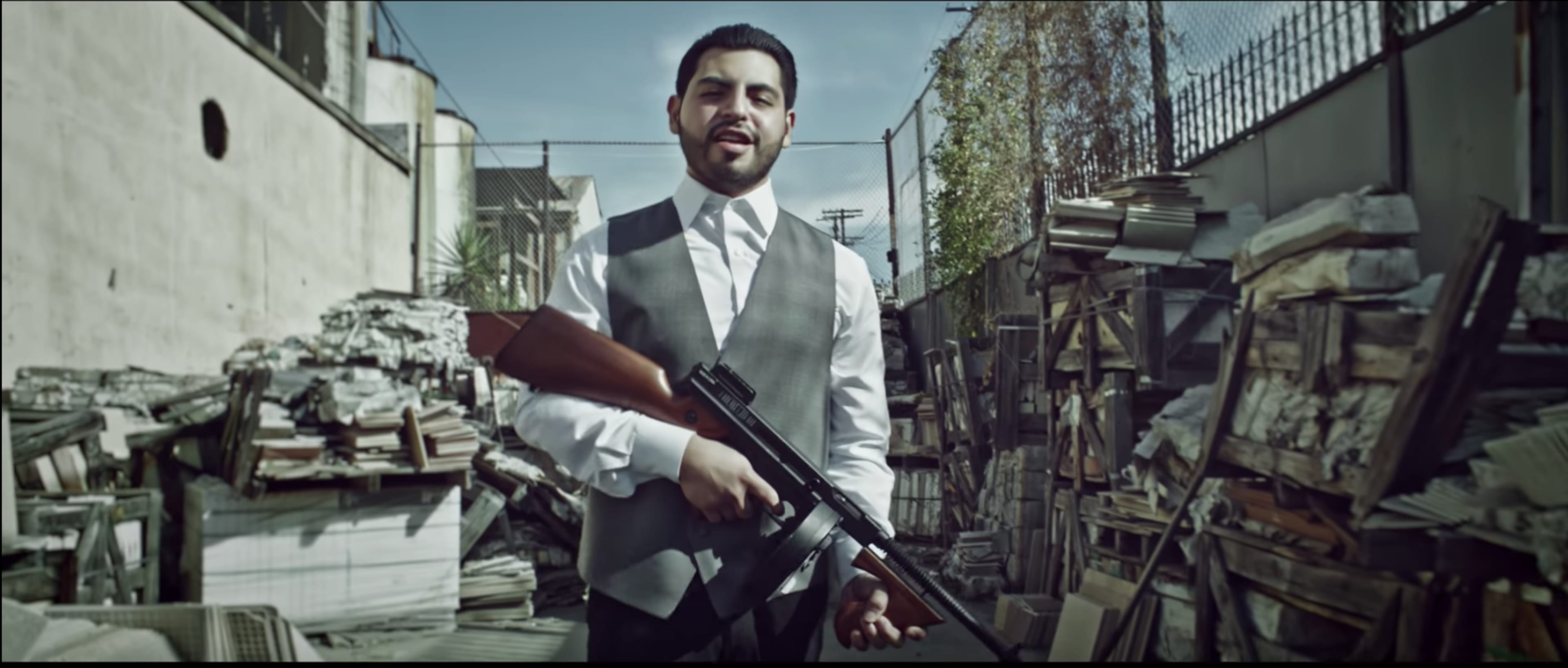A man in a suit holds a rifle while standing in a cluttered alley surrounded by stacks of debris.