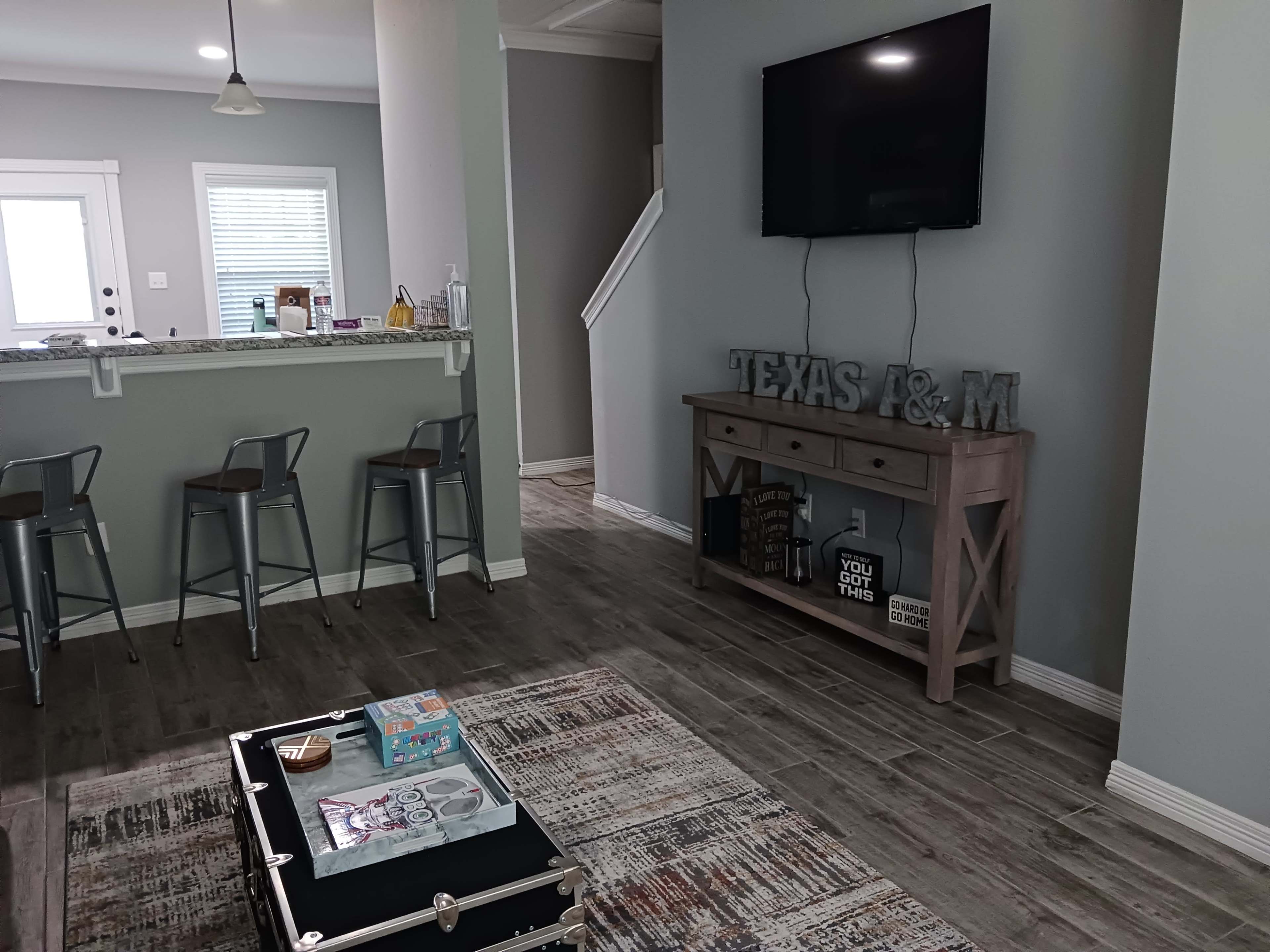 The image shows a modern living space with a gray wall, a television mounted on the wall, a kitchen bar with metal stools, and a decorative table featuring a rustic design with books and small items.