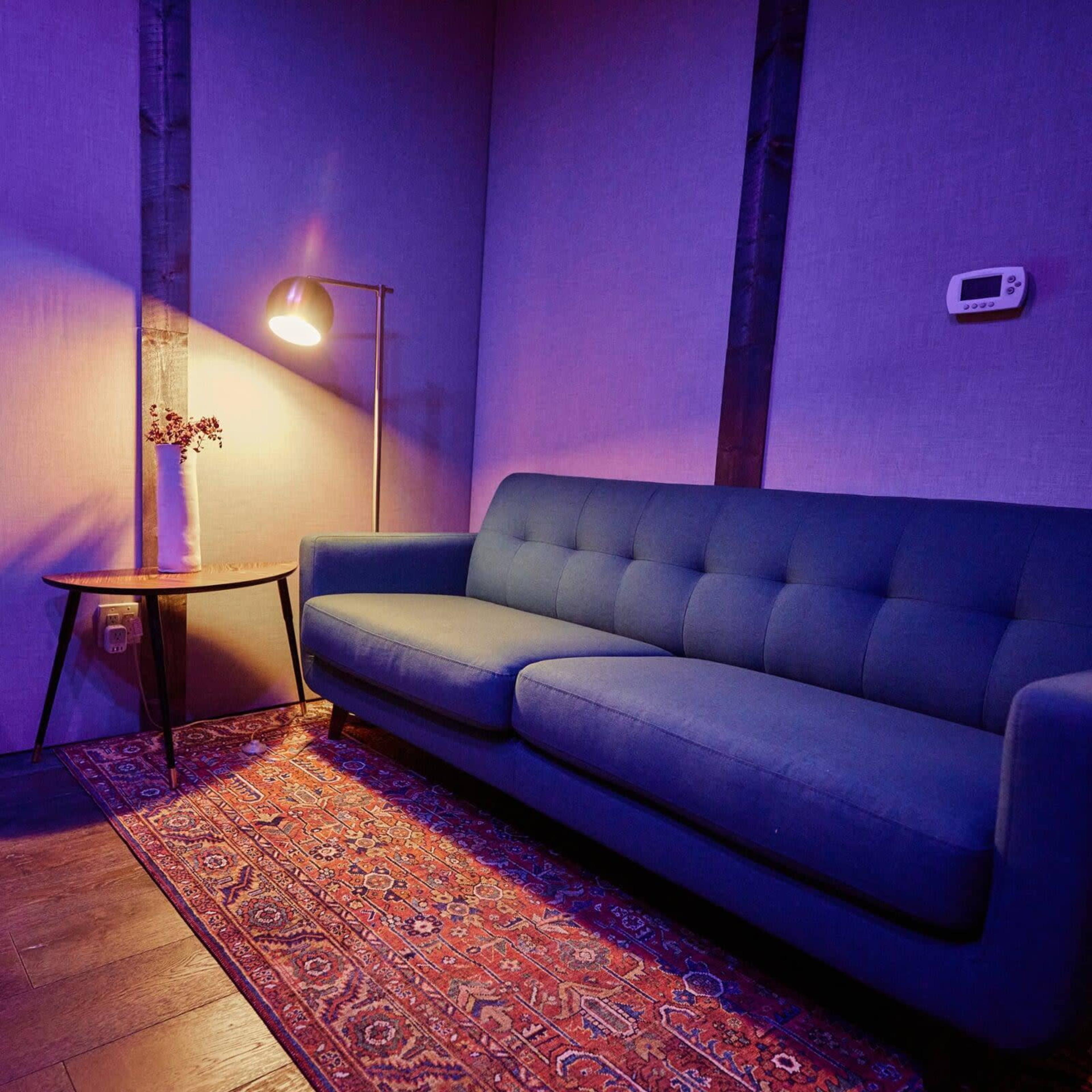 The image shows a modern living space with a blue couch, a small round table, a floor lamp, and a patterned rug on dark flooring.
