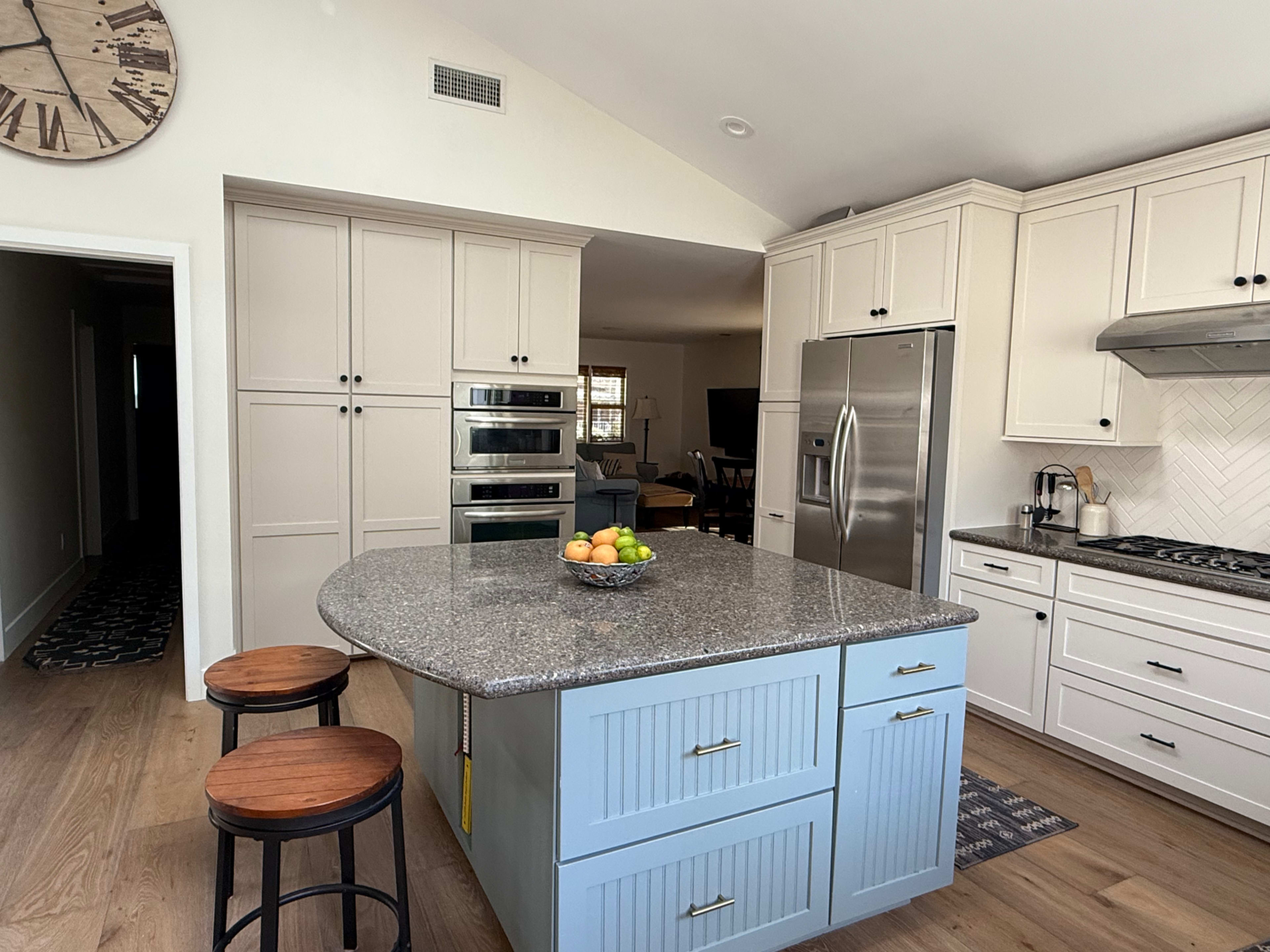 A modern kitchen features a large central island with a gray countertop and a bowl of fruit, surrounded by white cabinetry and stainless steel appliances.