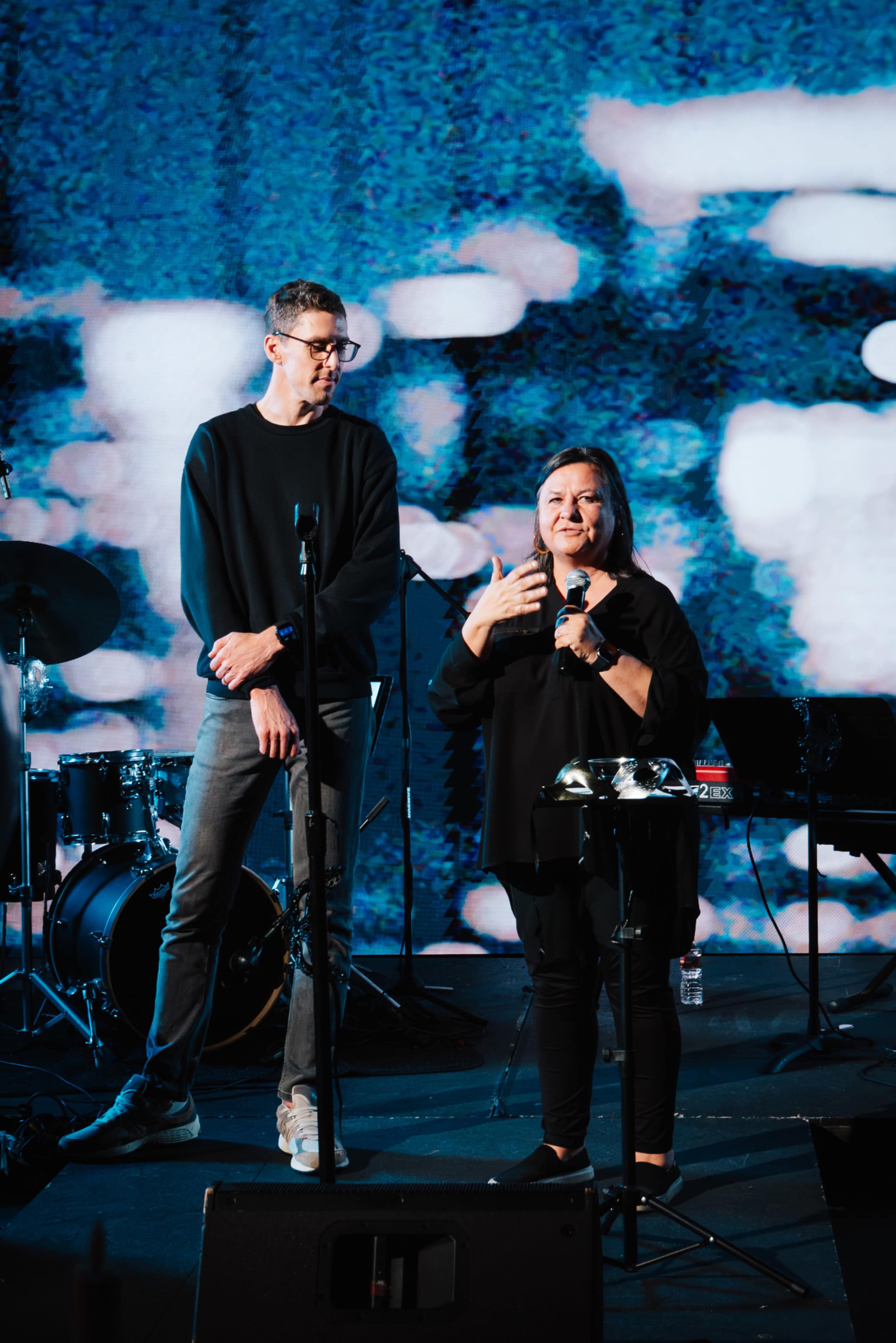 A man and a woman are speaking on stage in front of a large screen, with musical instruments visible in the background.