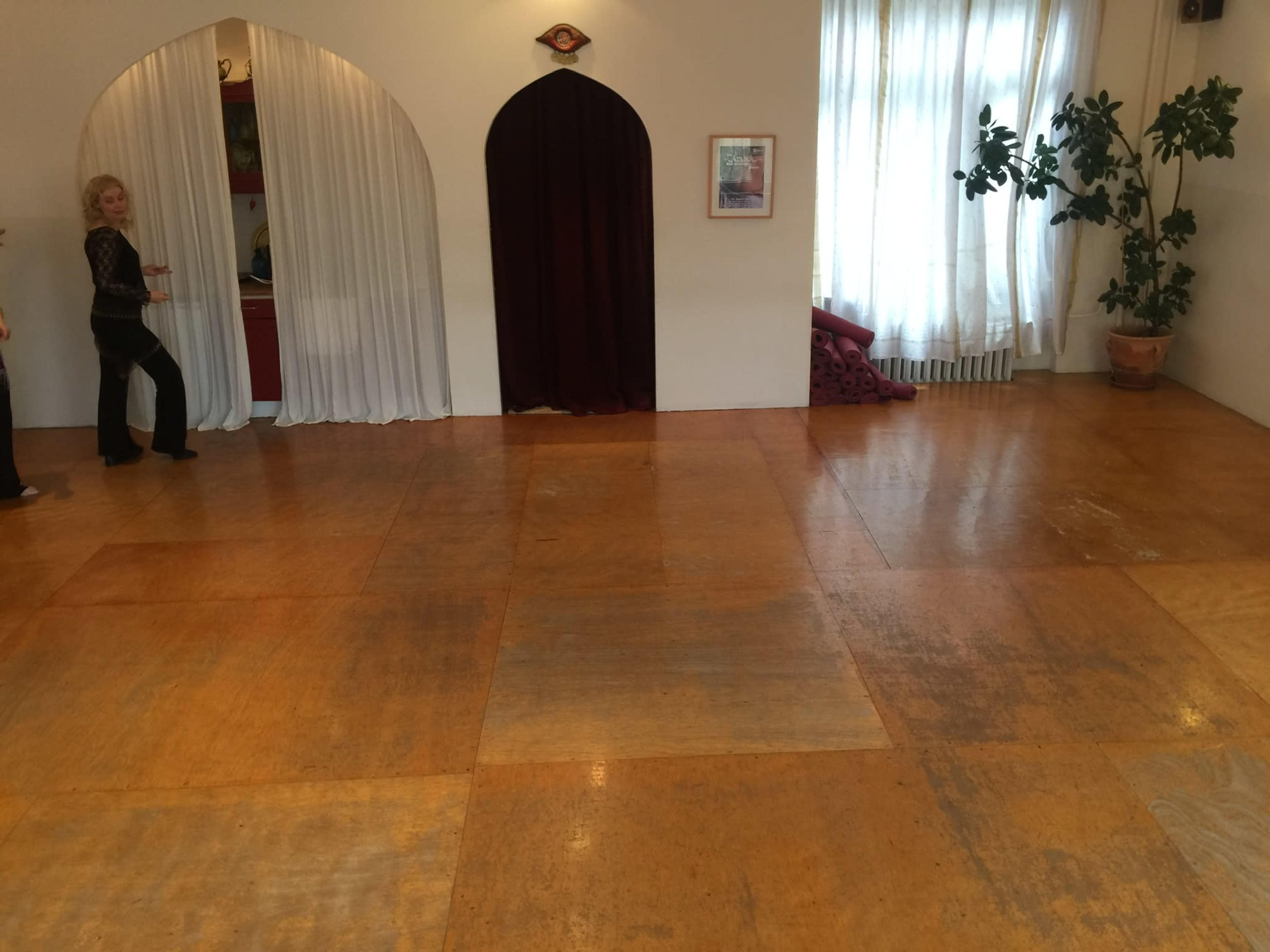A spacious room features an arched doorway, a white curtain, and a large window with a potted plant, alongside a polished wooden floor.