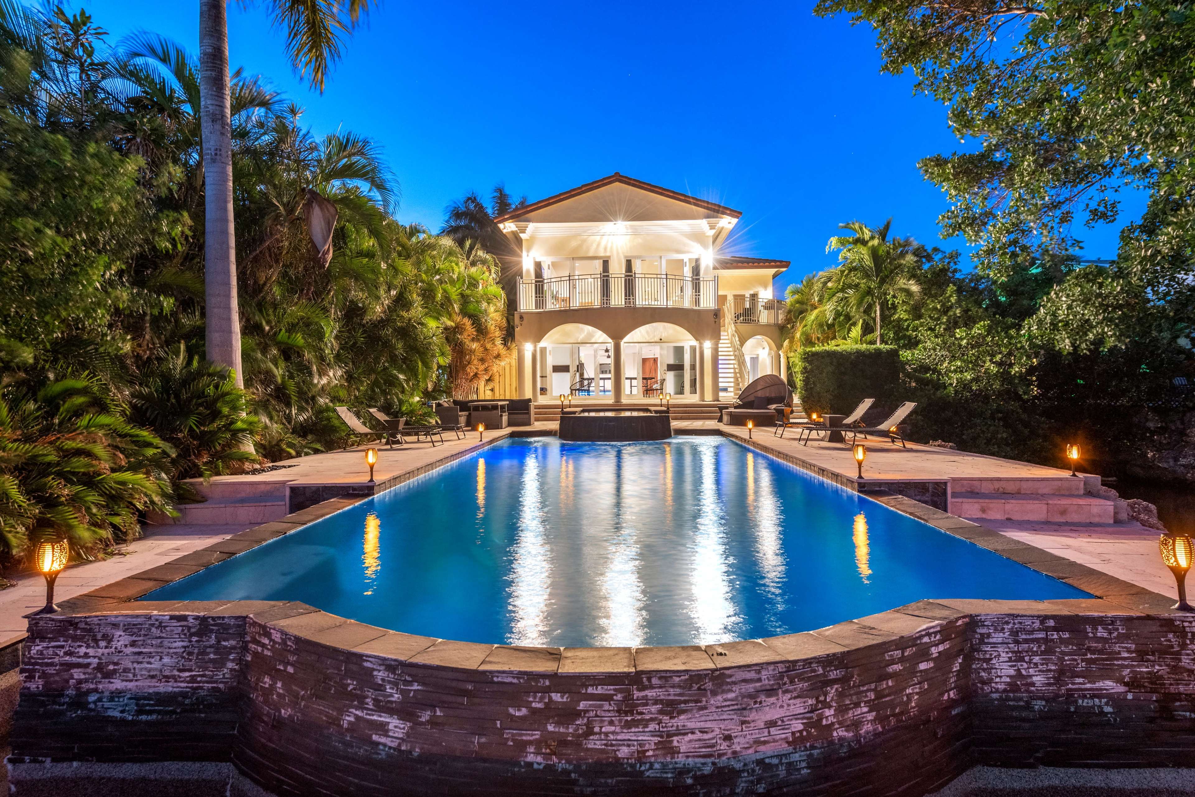 A large house with a balcony overlooks a rectangular swimming pool surrounded by tropical vegetation and lounge chairs.