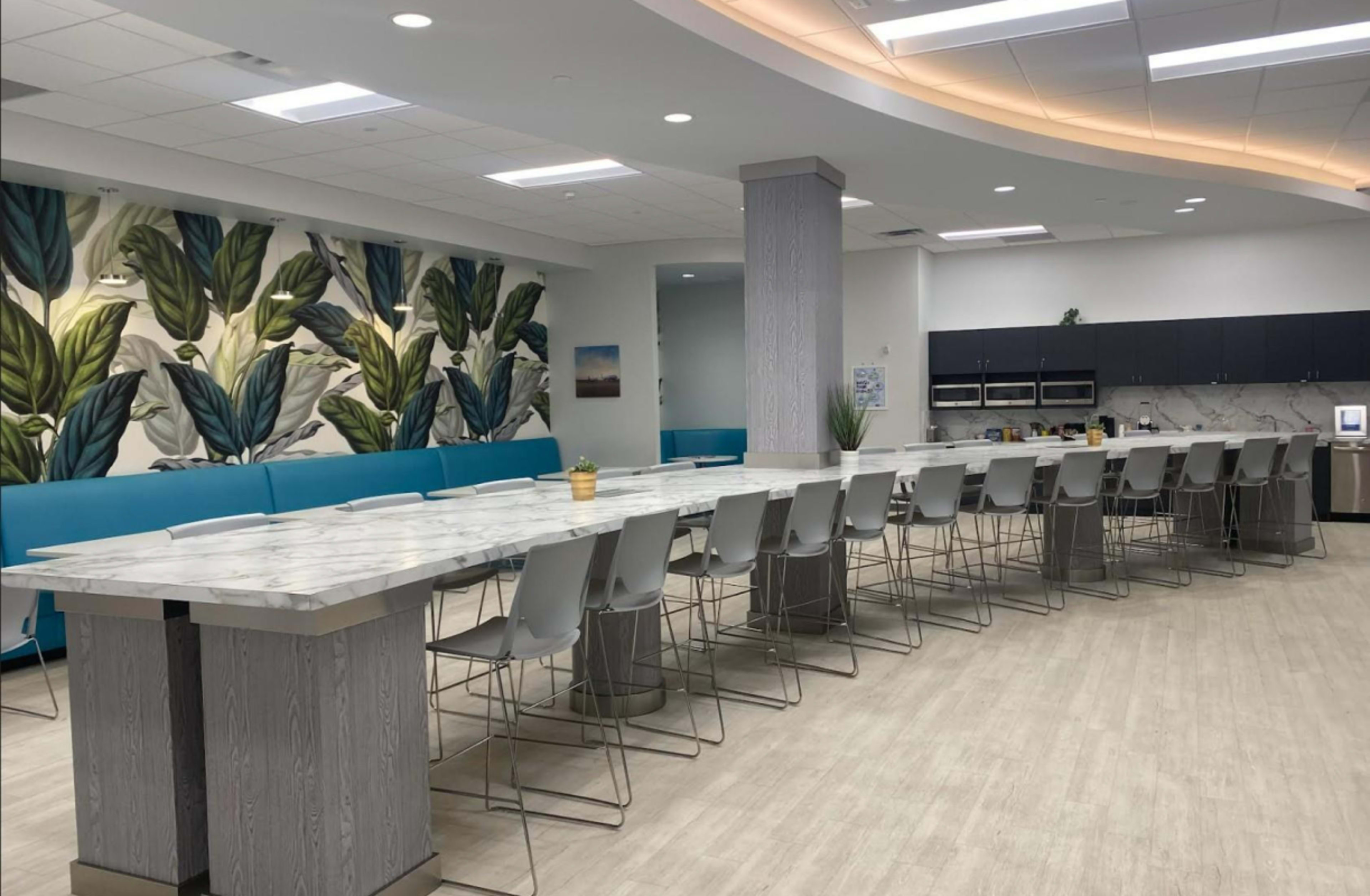A modern kitchen area with a long marble-topped table surrounded by sleek gray chairs, complemented by leafy wallpaper and blue seating along one wall.