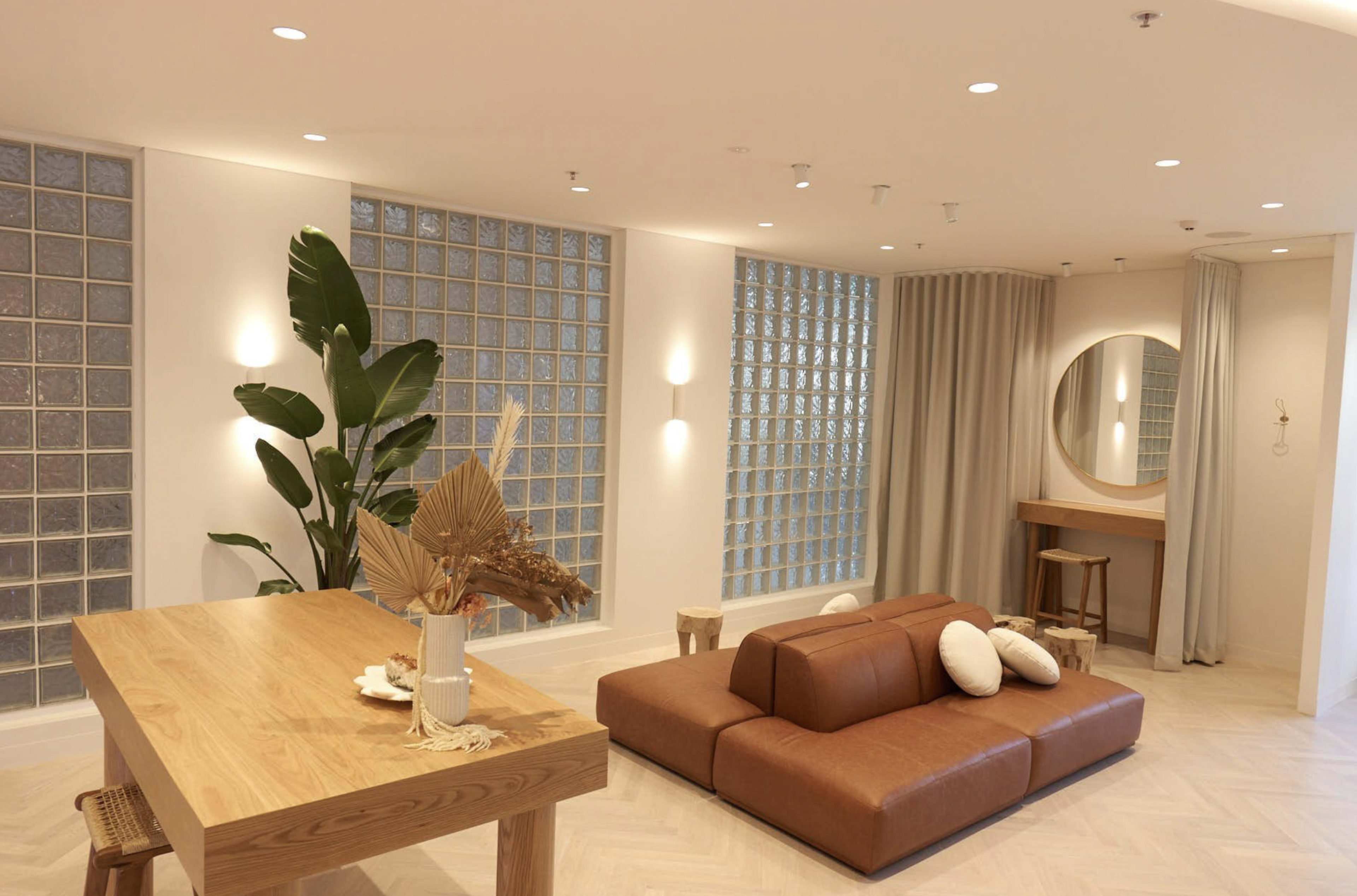 A modern interior space featuring a brown leather sofa, a wooden dining table, large glass block windows, and a circular mirror on the wall.