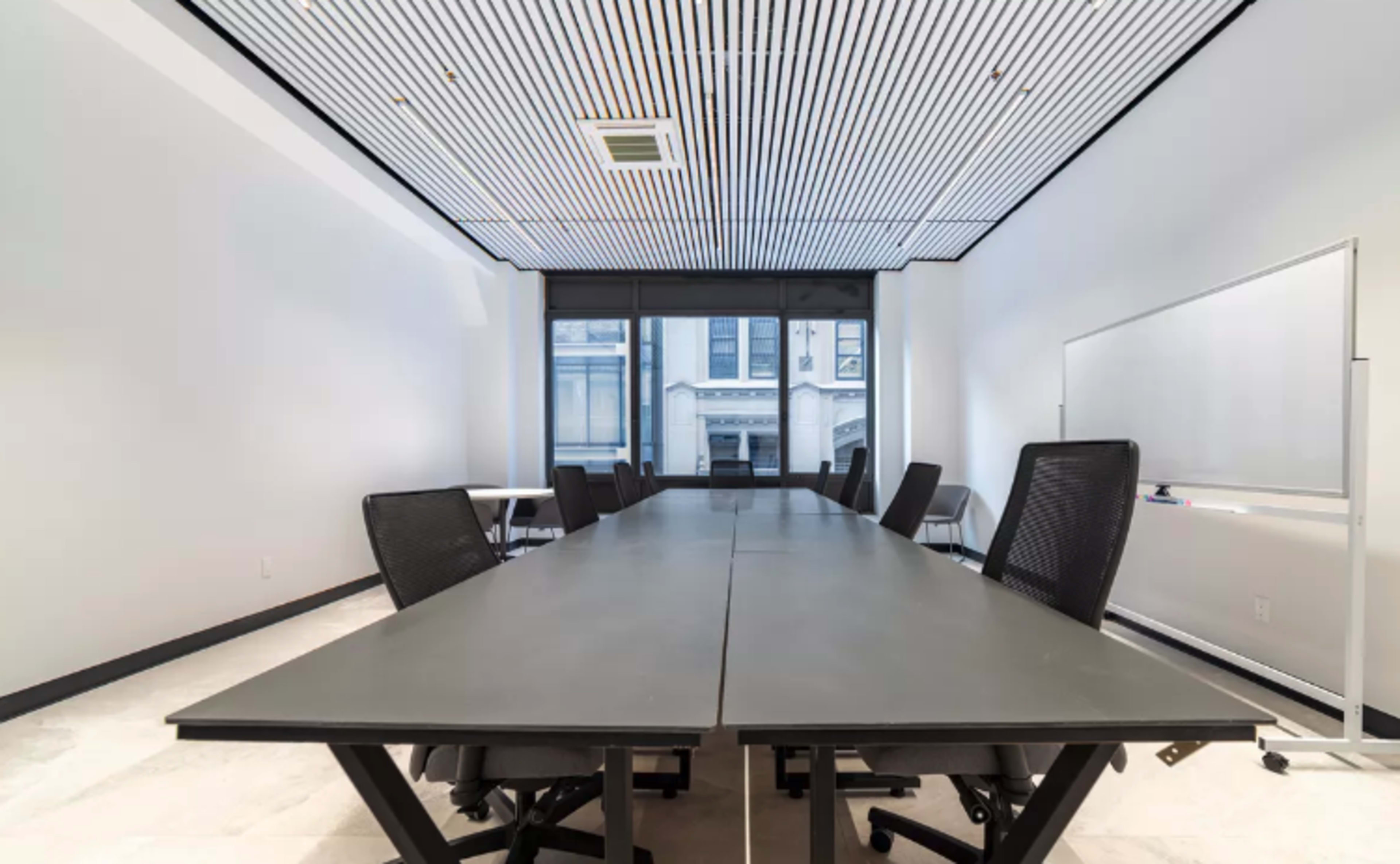 A long conference table with chairs is centered in a minimalist meeting room featuring large windows.
