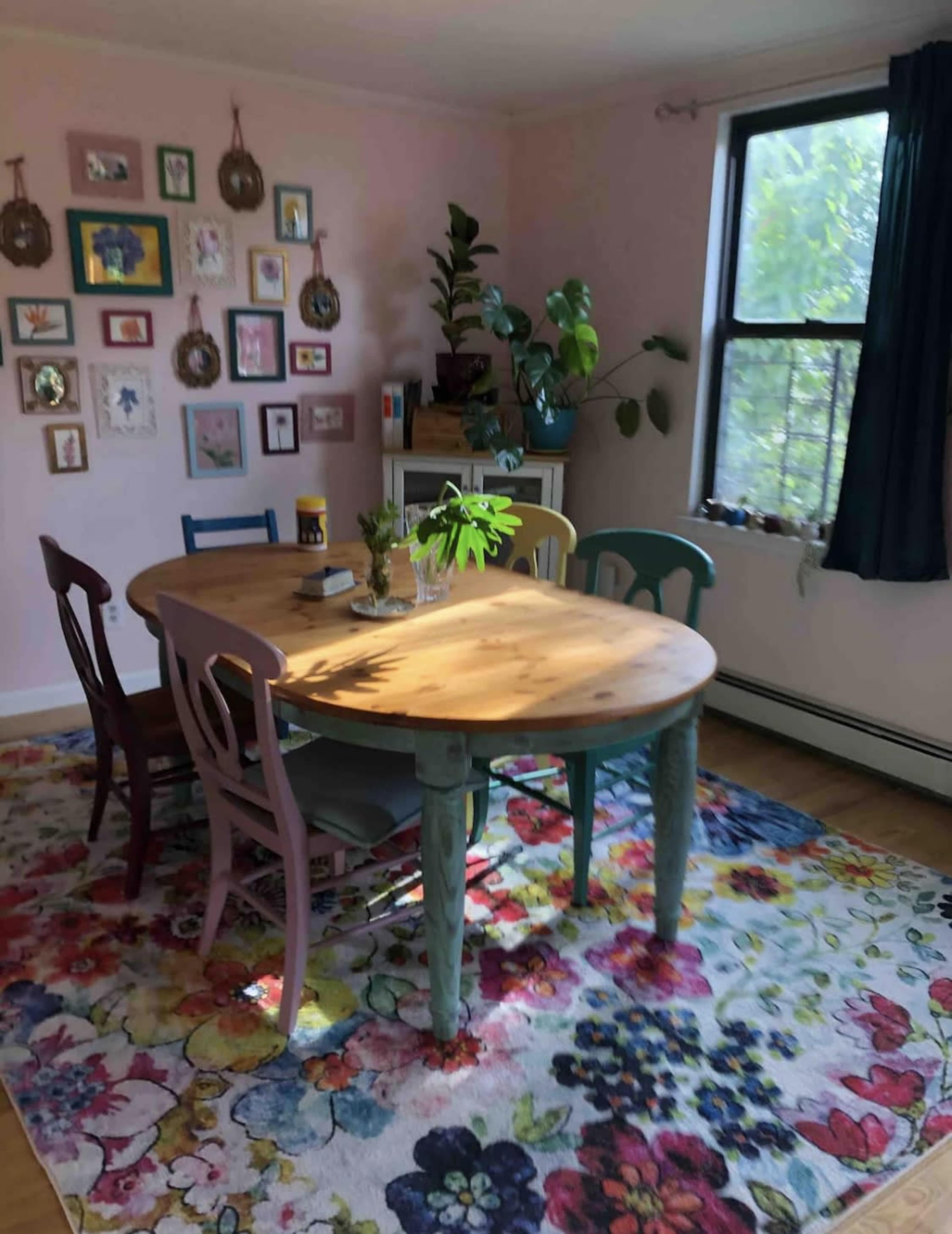 A round wooden table surrounded by colorful chairs is set on a floral rug in a room adorned with framed pictures on the wall.