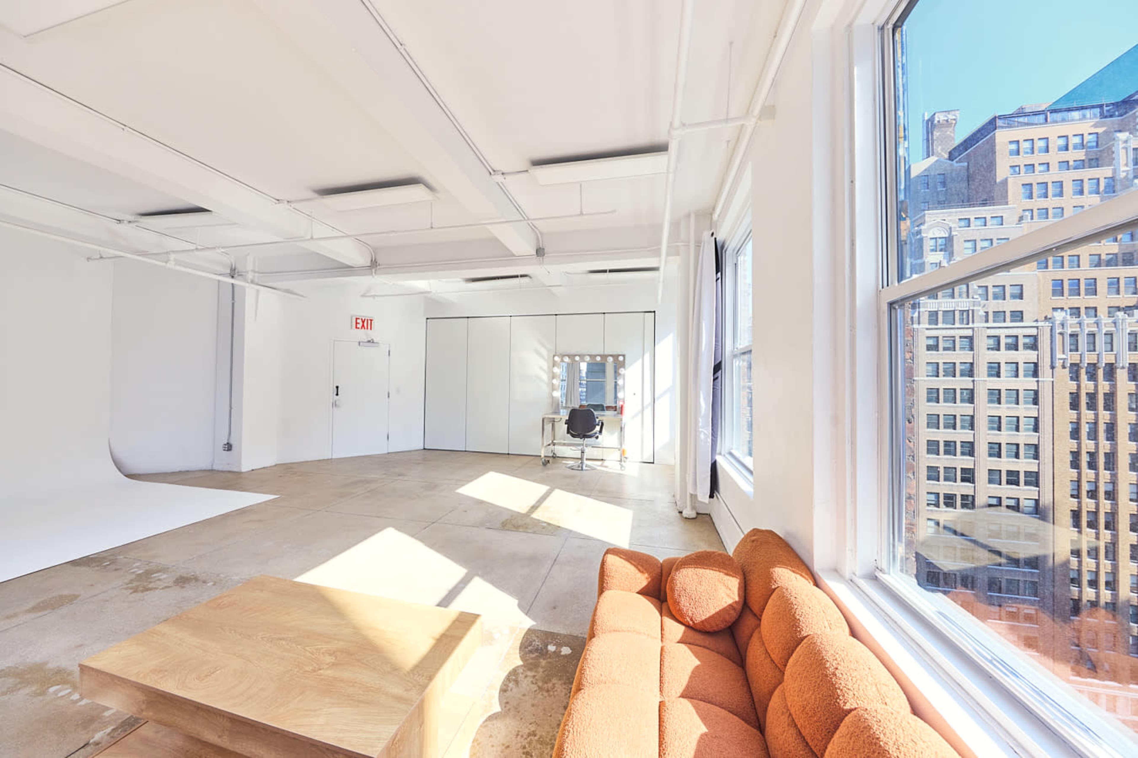 A bright, spacious room features large windows, a cozy orange couch, a wooden table, and a desk with a chair.