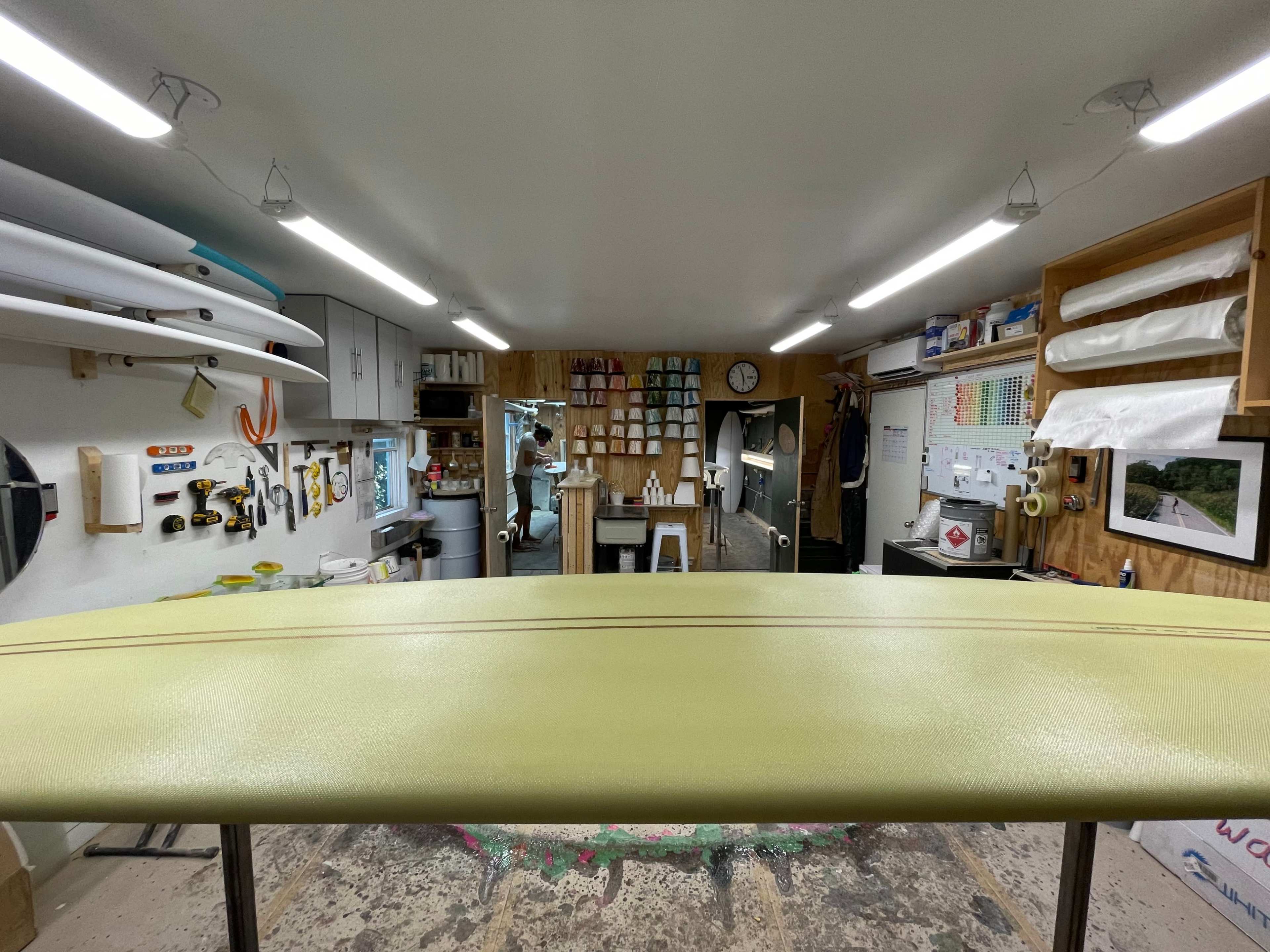 The image shows a surfboard shaping workshop with tools, materials, and a finished surfboard displayed prominently in the foreground.