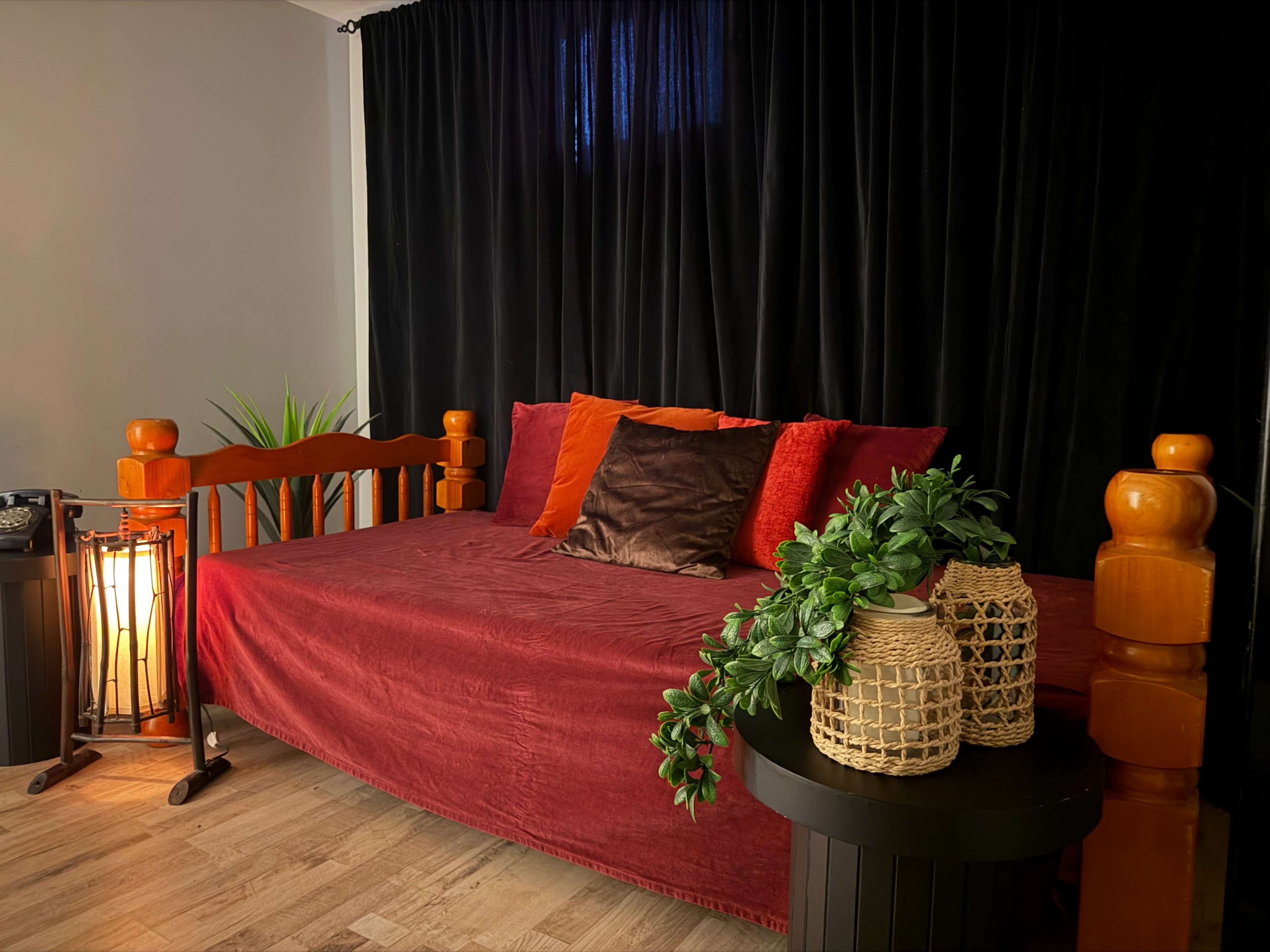The scene features a bed with red and orange pillows against a black curtain, accompanied by a woven plant holder and a small lamp.