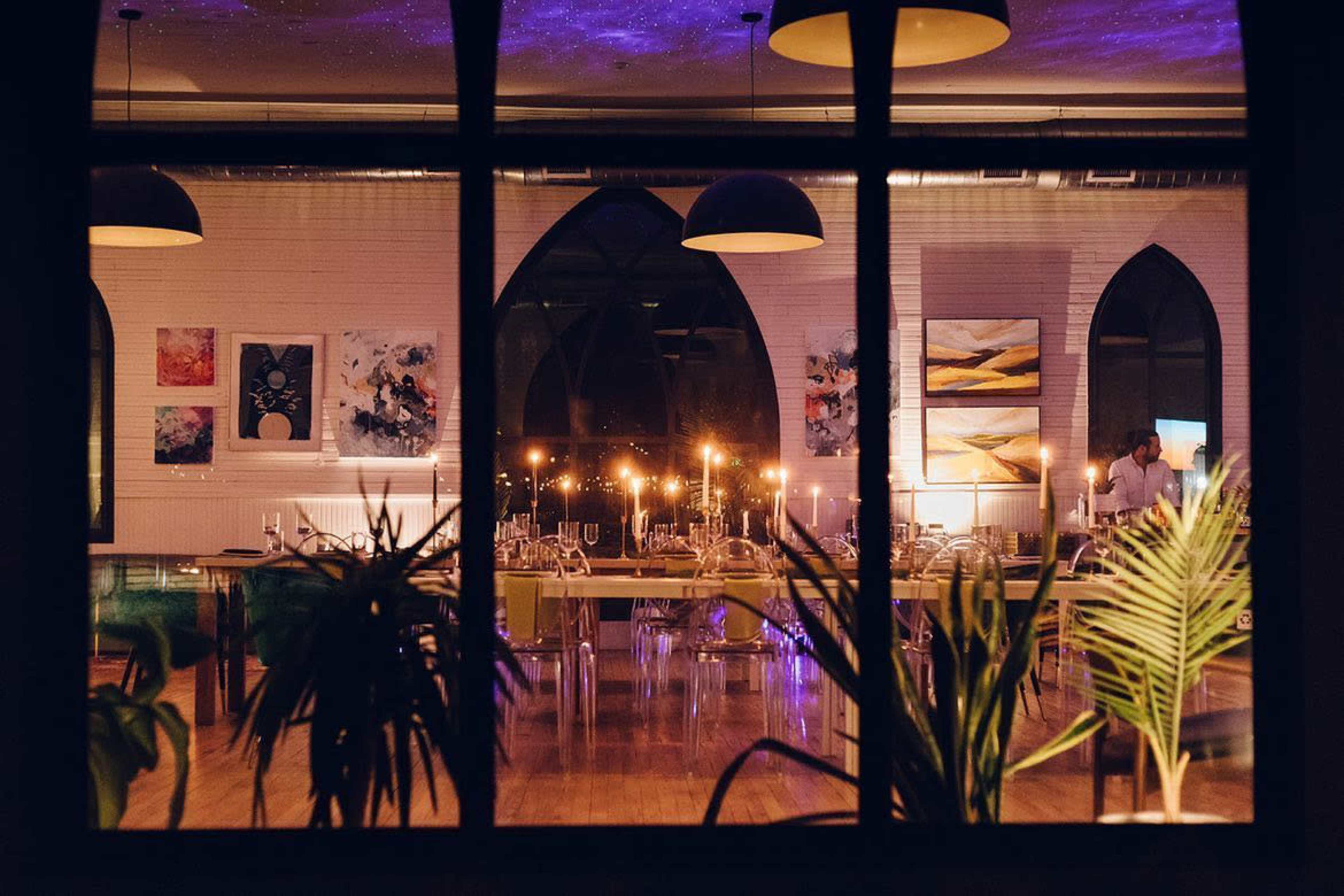 The image shows a well-lit restaurant interior visible through a large window, featuring elegant tables set with candles and artwork on the walls.