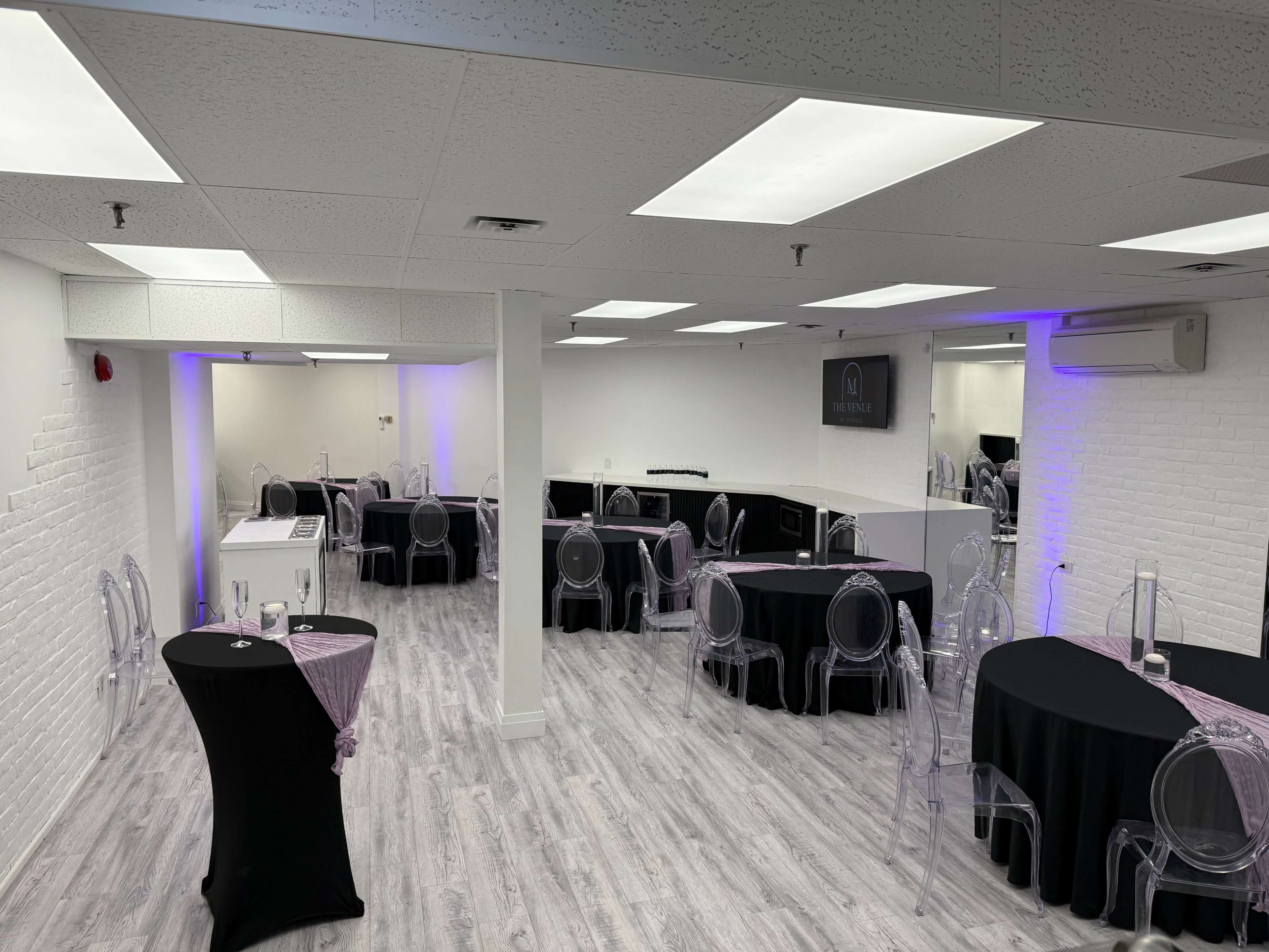 The image shows a spacious, well-lit event venue with several tables set up, featuring a mix of black and clear chairs.
