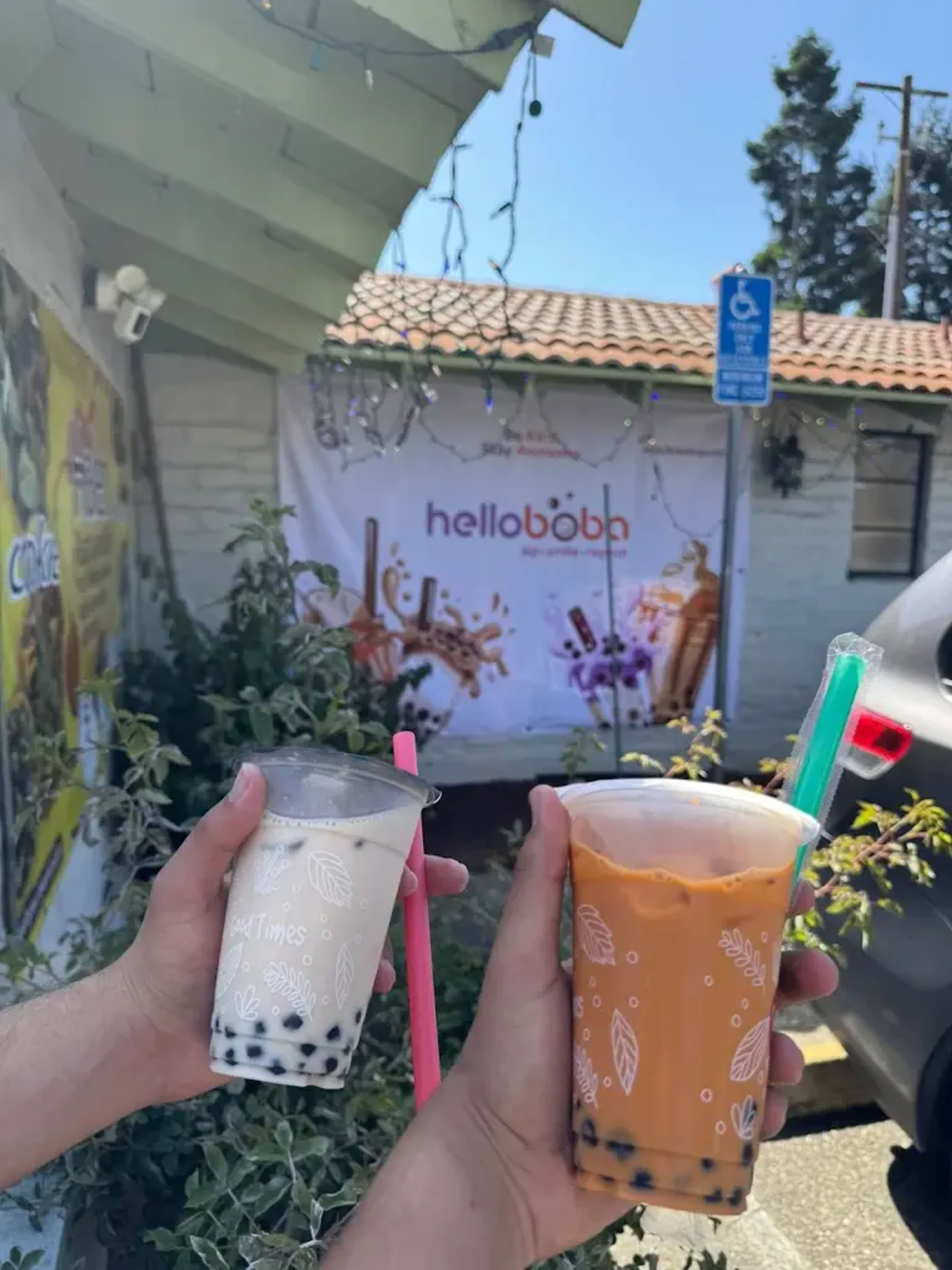 Two hands hold bubble tea drinks, one with a cream-colored beverage and the other with an orange drink, in front of a "helloboba" banner.