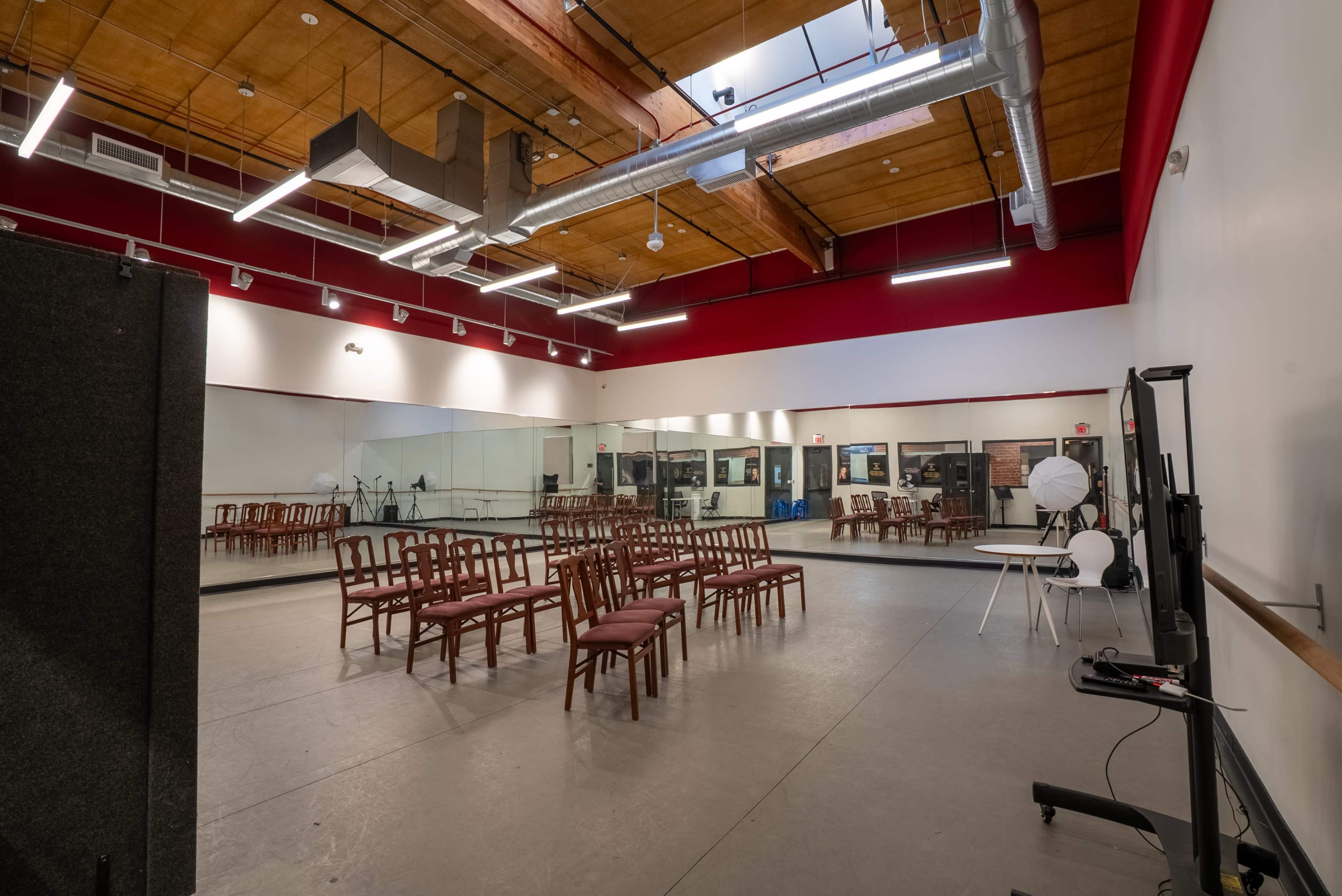 The image shows a spacious rehearsal studio with a mirrored wall, several wooden chairs arranged in rows, and various lighting equipment set up.