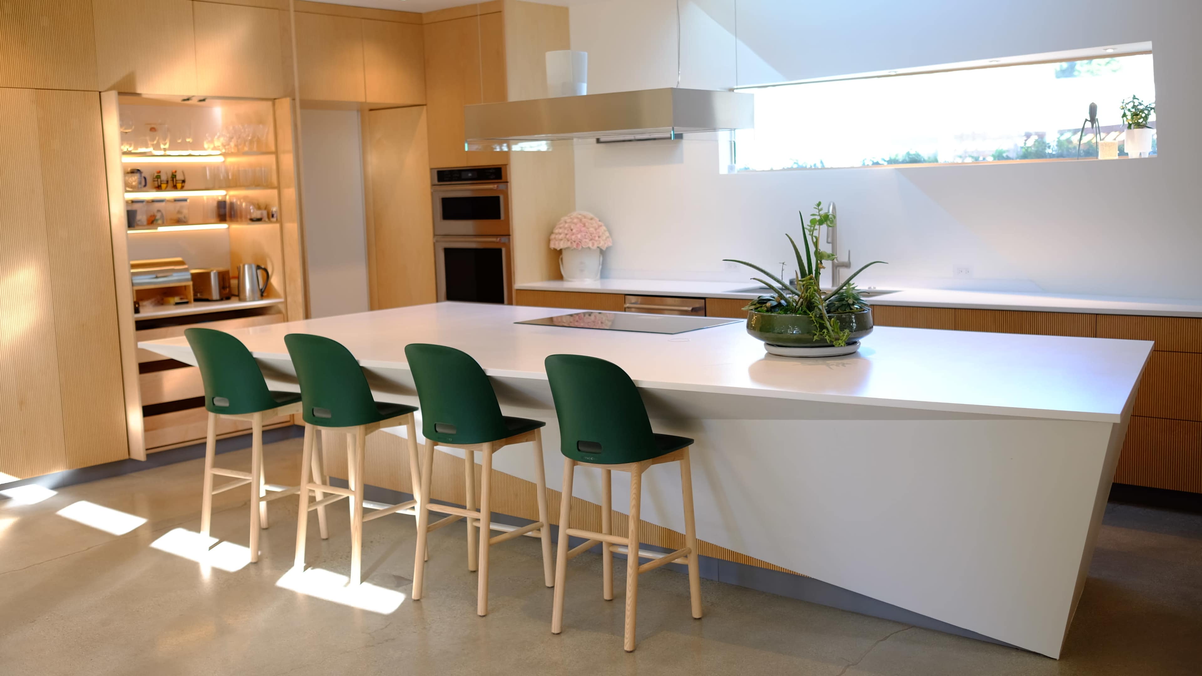 The image shows a modern kitchen with a large white island, four green stools, and wooden cabinetry.