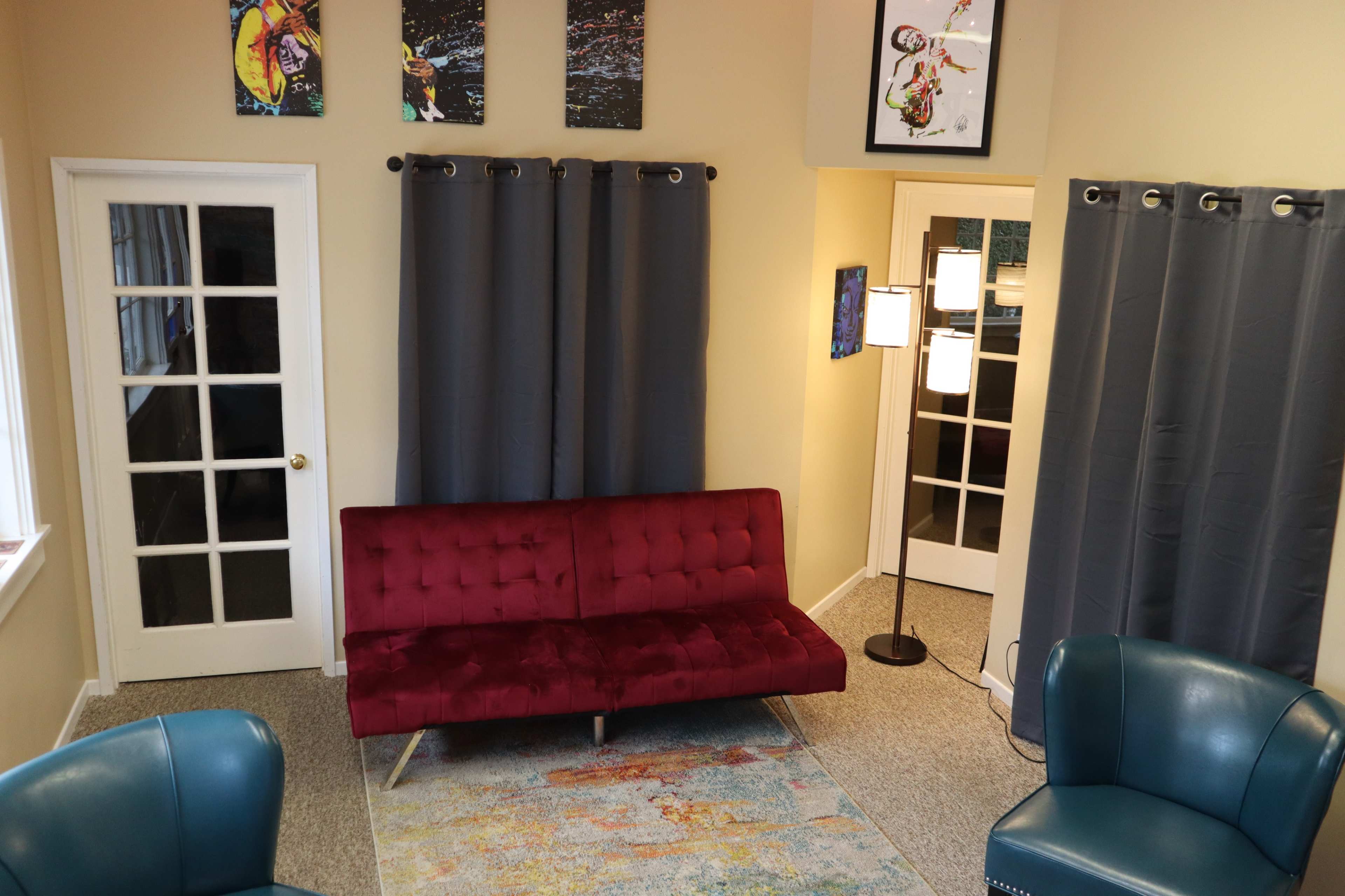 The room features a red velvet couch, two teal chairs, gray curtains, and artwork on the walls, with a floor lamp in one corner.