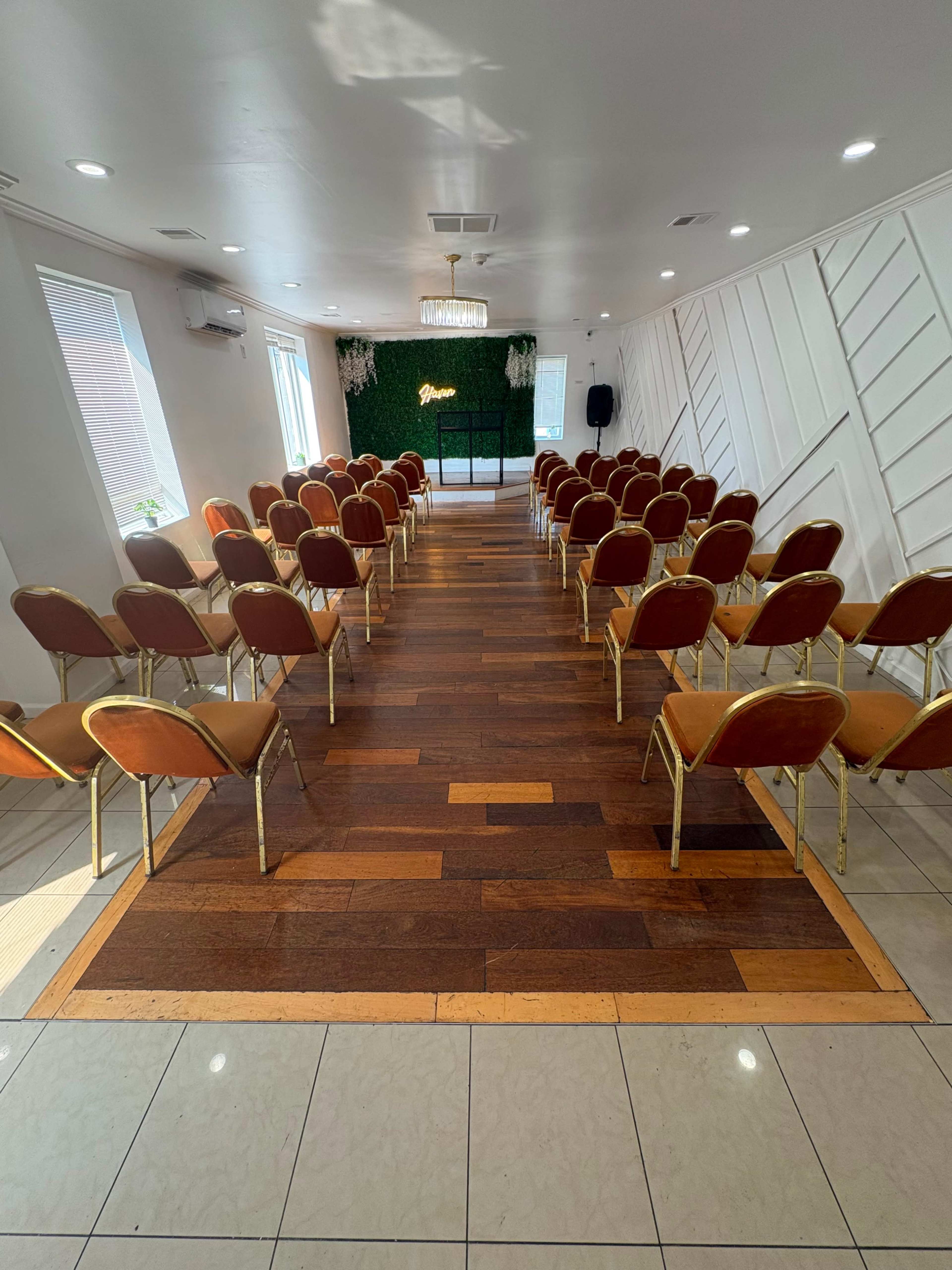 The image shows a well-arranged indoor venue with rows of empty chairs facing a small stage adorned with greenery.