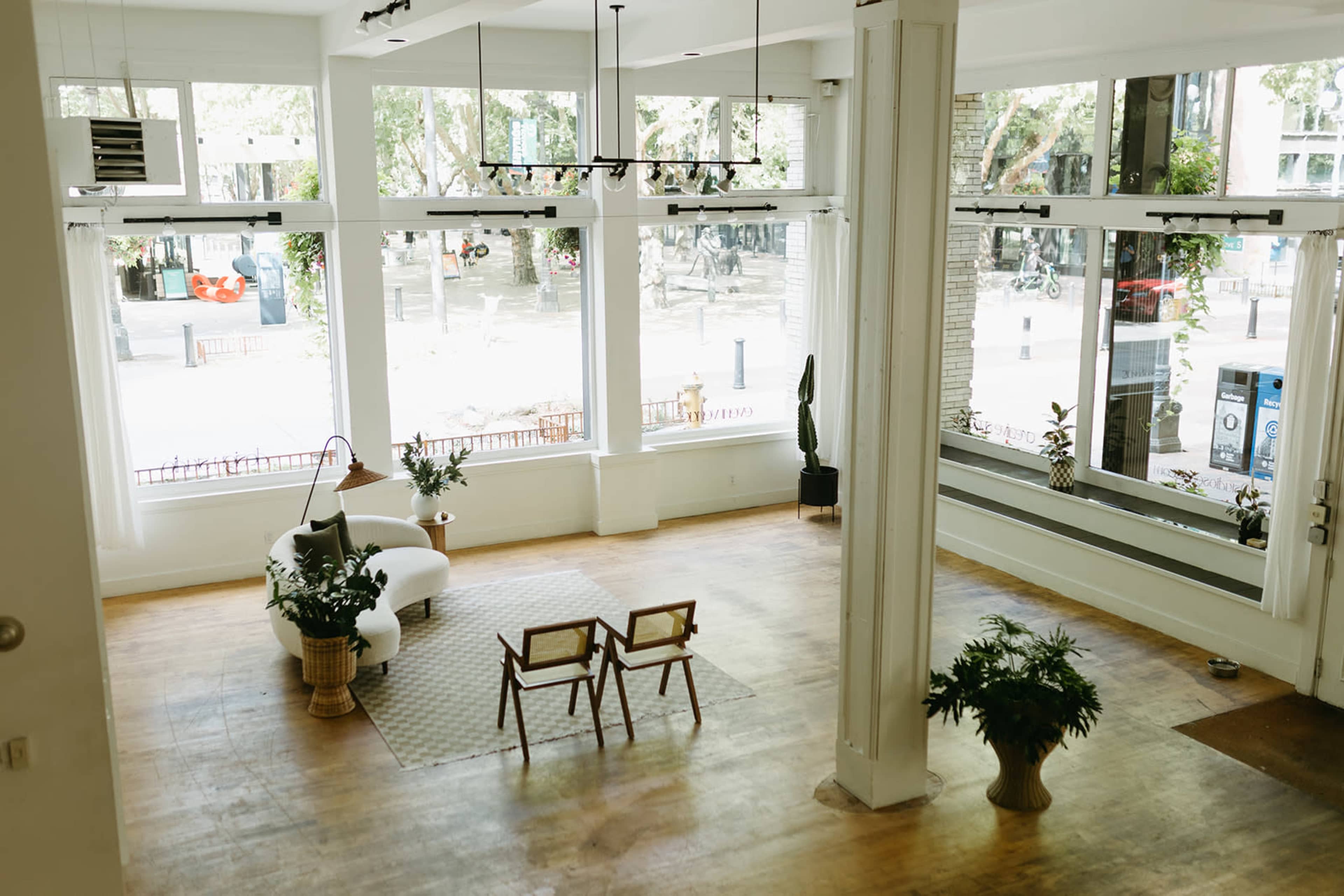 The image shows a bright, open space with large windows, light wooden flooring, and minimal furniture, including a sofa and two chairs positioned near indoor plants.