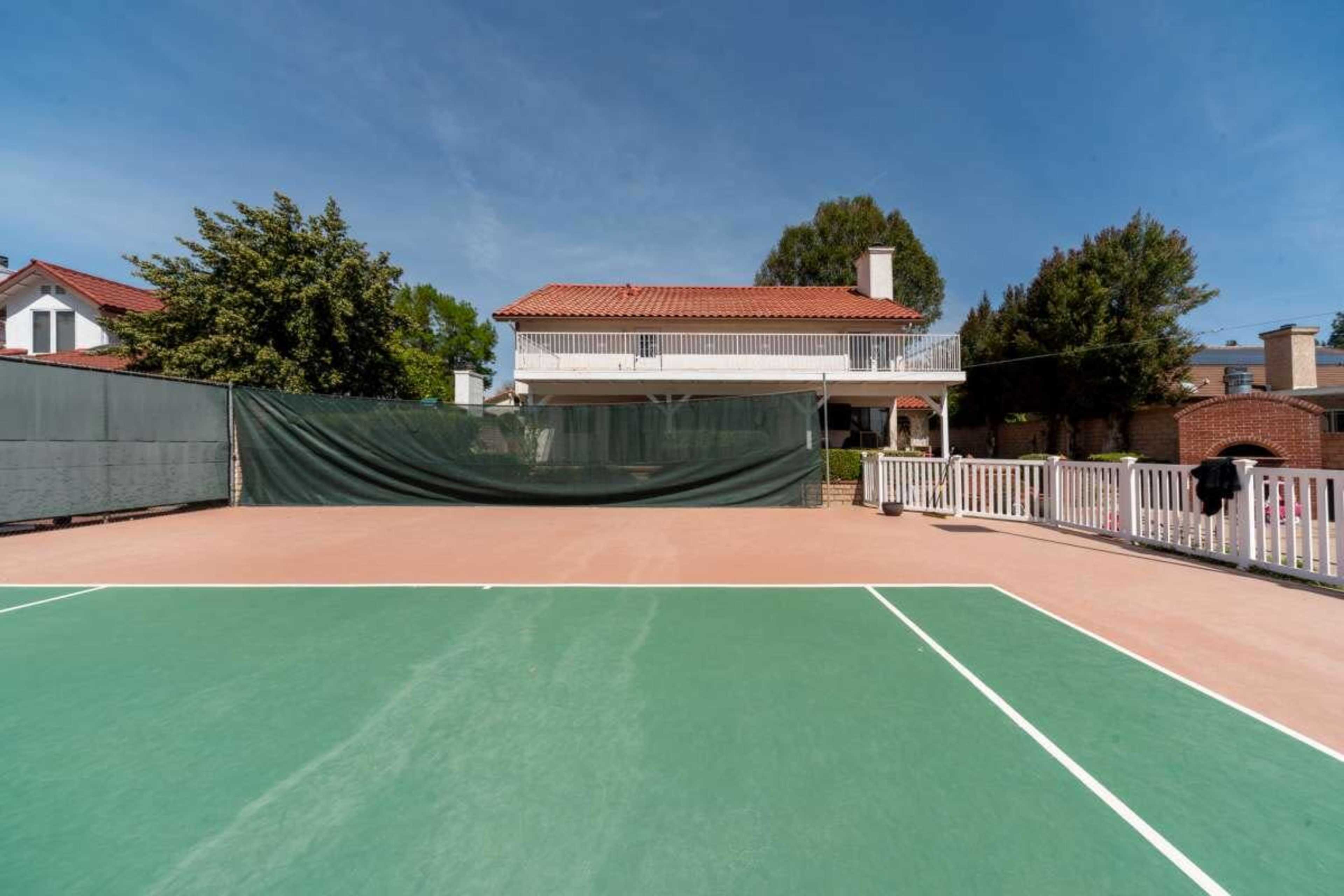 A tennis court with a green surface is shown in the foreground, and a two-story house with a red-tiled roof is visible in the background.