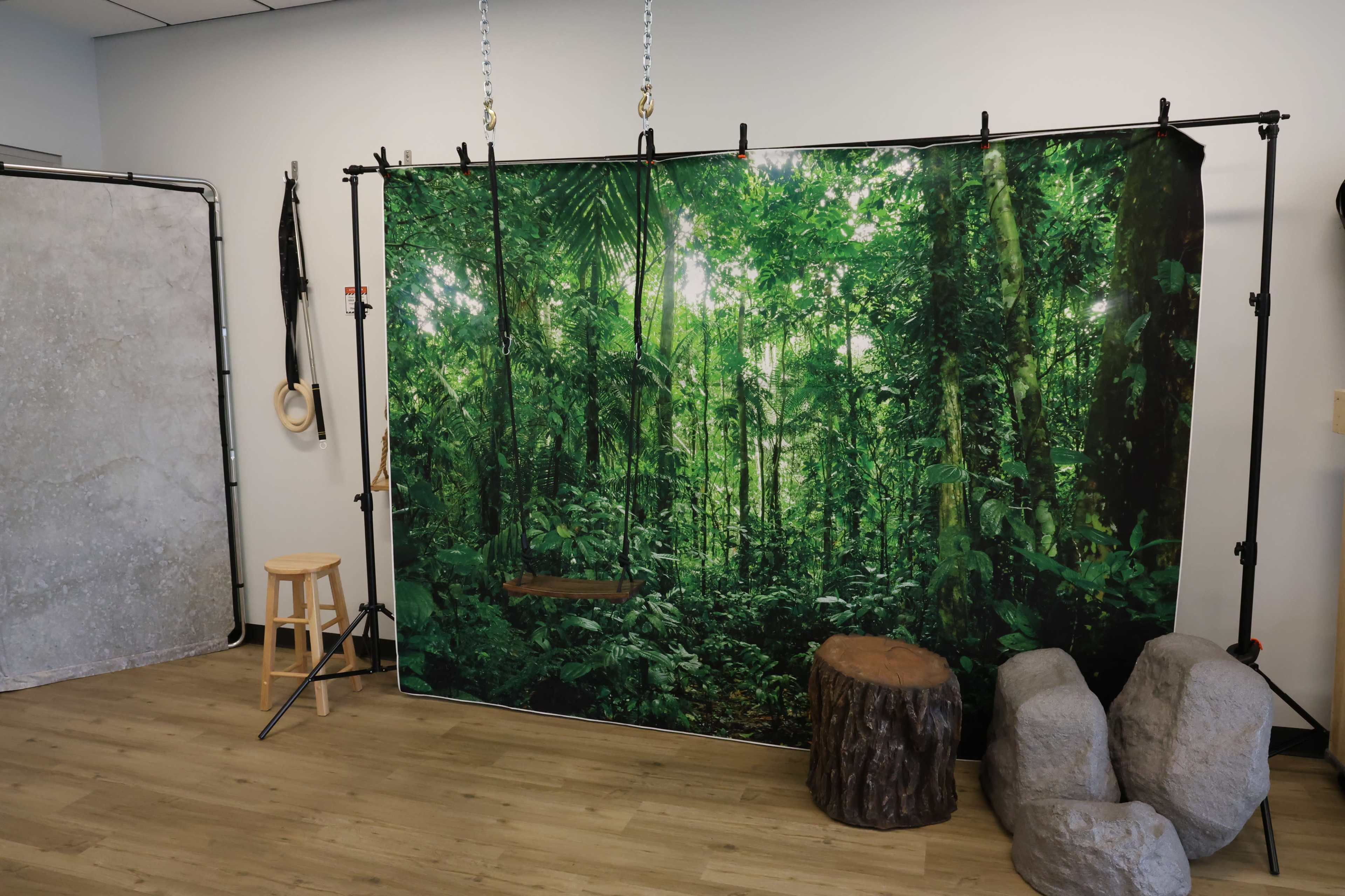 A backdrop of a dense green jungle with a swing, accompanied by a wooden stool and stone props in a studio setting.