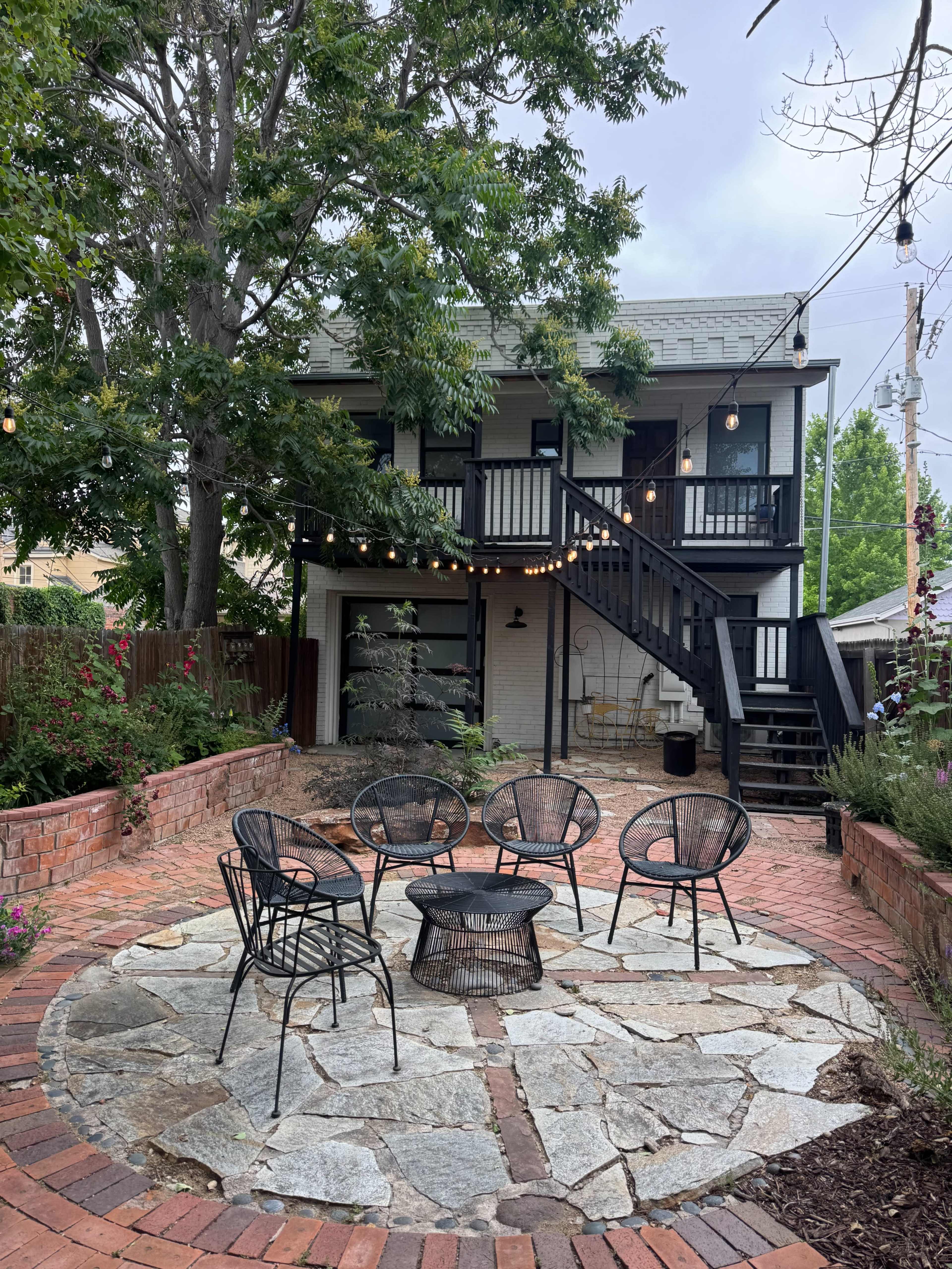 The image shows a backyard with a stone and brick patio featuring five black chairs arranged around a circular fire pit, in front of a two-story house with a staircase and string lights overhead.
