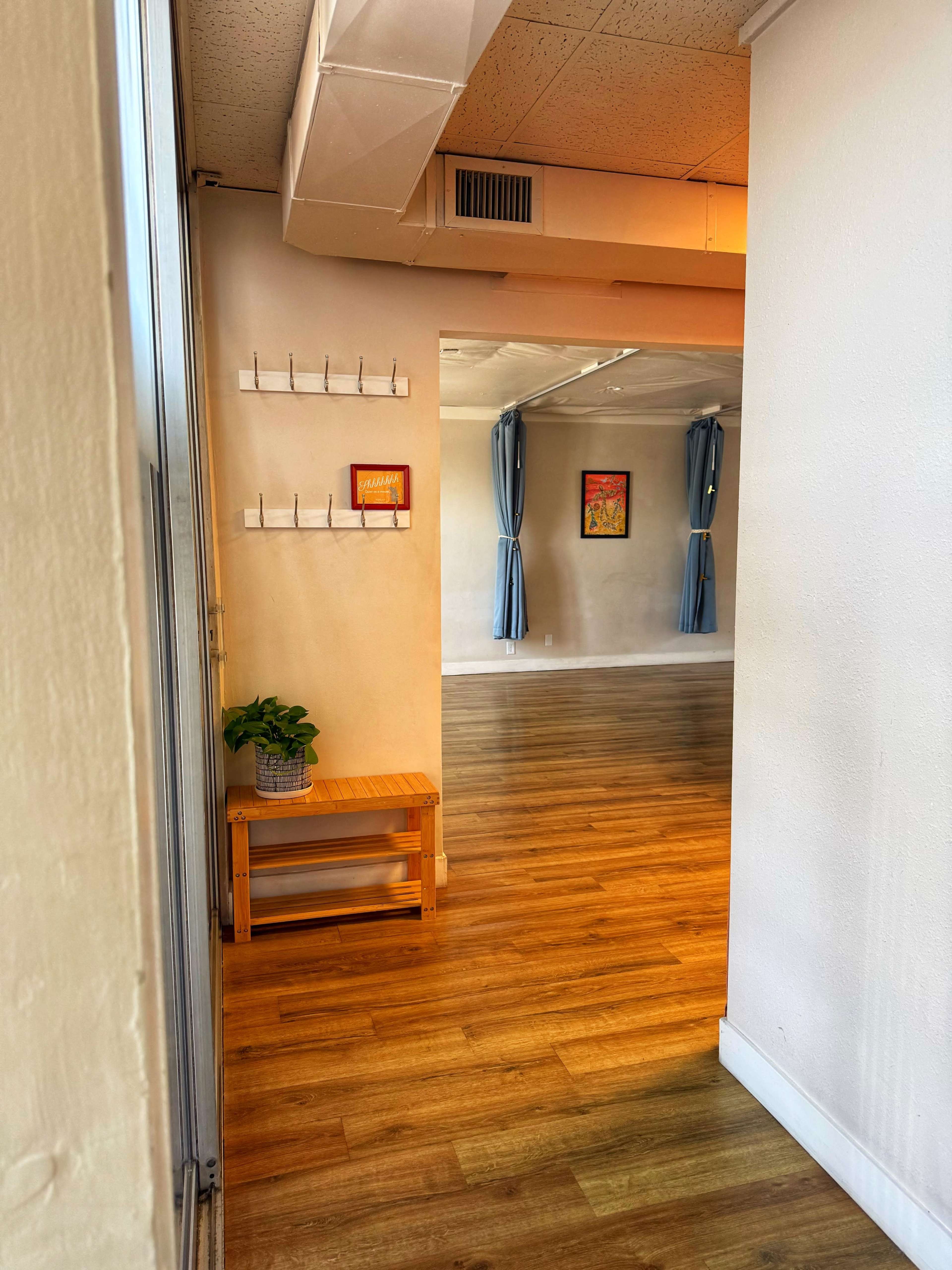 The image shows a hallway leading to a spacious, well-lit room with wooden flooring and blue curtains.