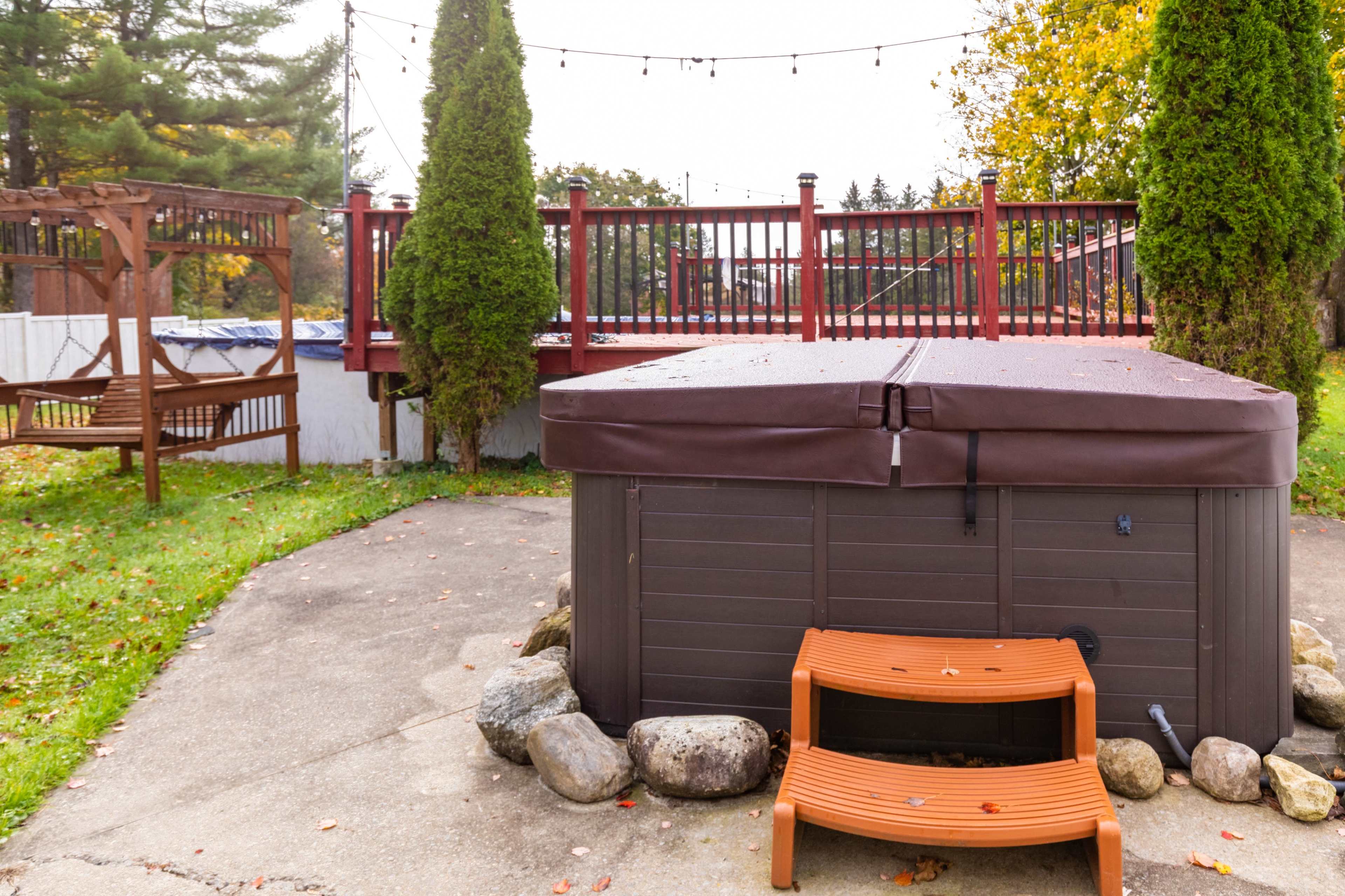 A hot tub sits on a paved area surrounded by landscaped grass and stones, with a deck featuring a railing and string lights in the background.