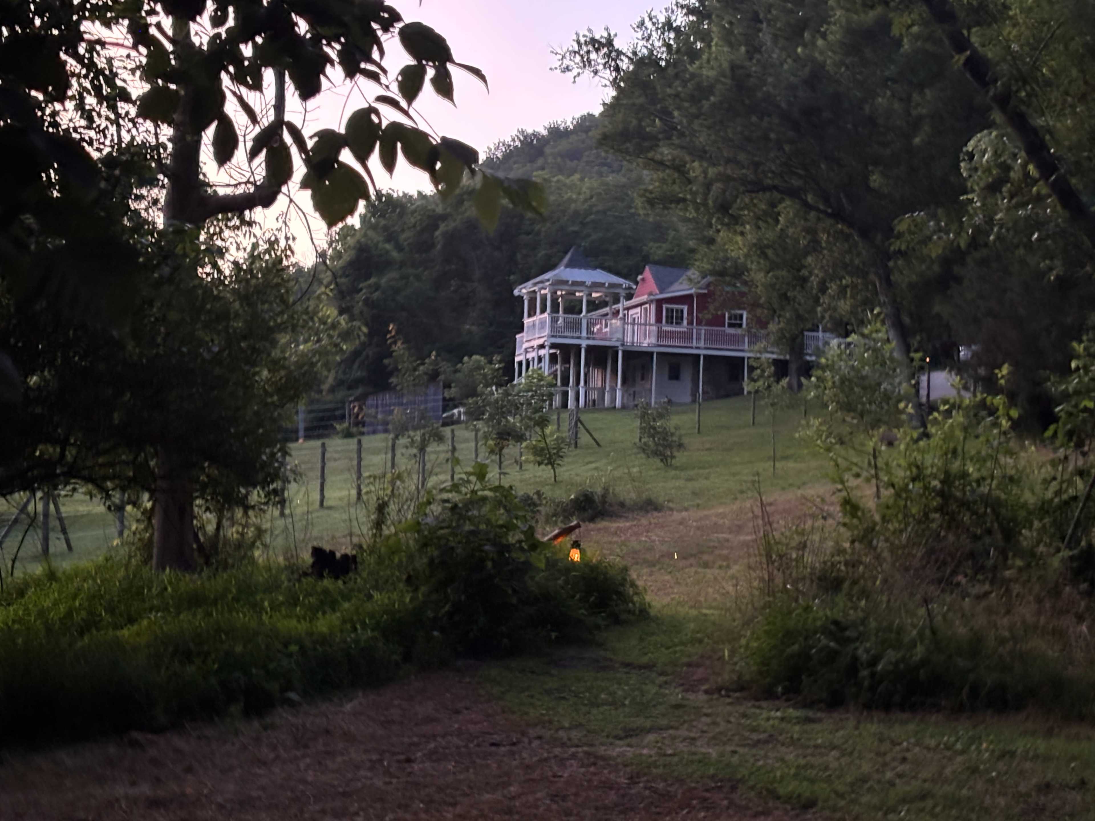 A large, two-story house with a wraparound porch is situated on a grassy hillside, surrounded by trees and a fenced area.