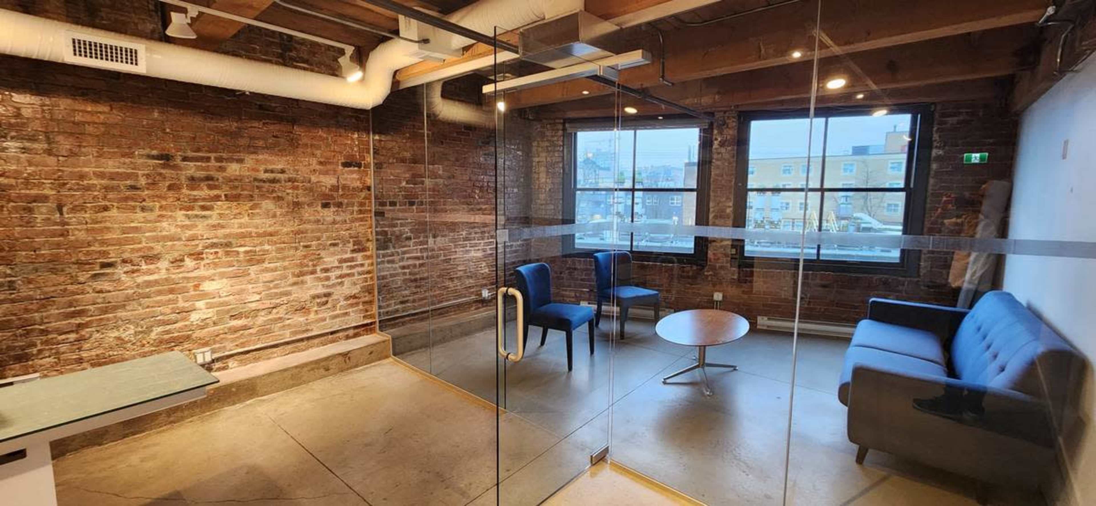 The image shows a modern office space featuring a glass meeting room with exposed brick walls, a sofa, and two chairs arranged around a small table.