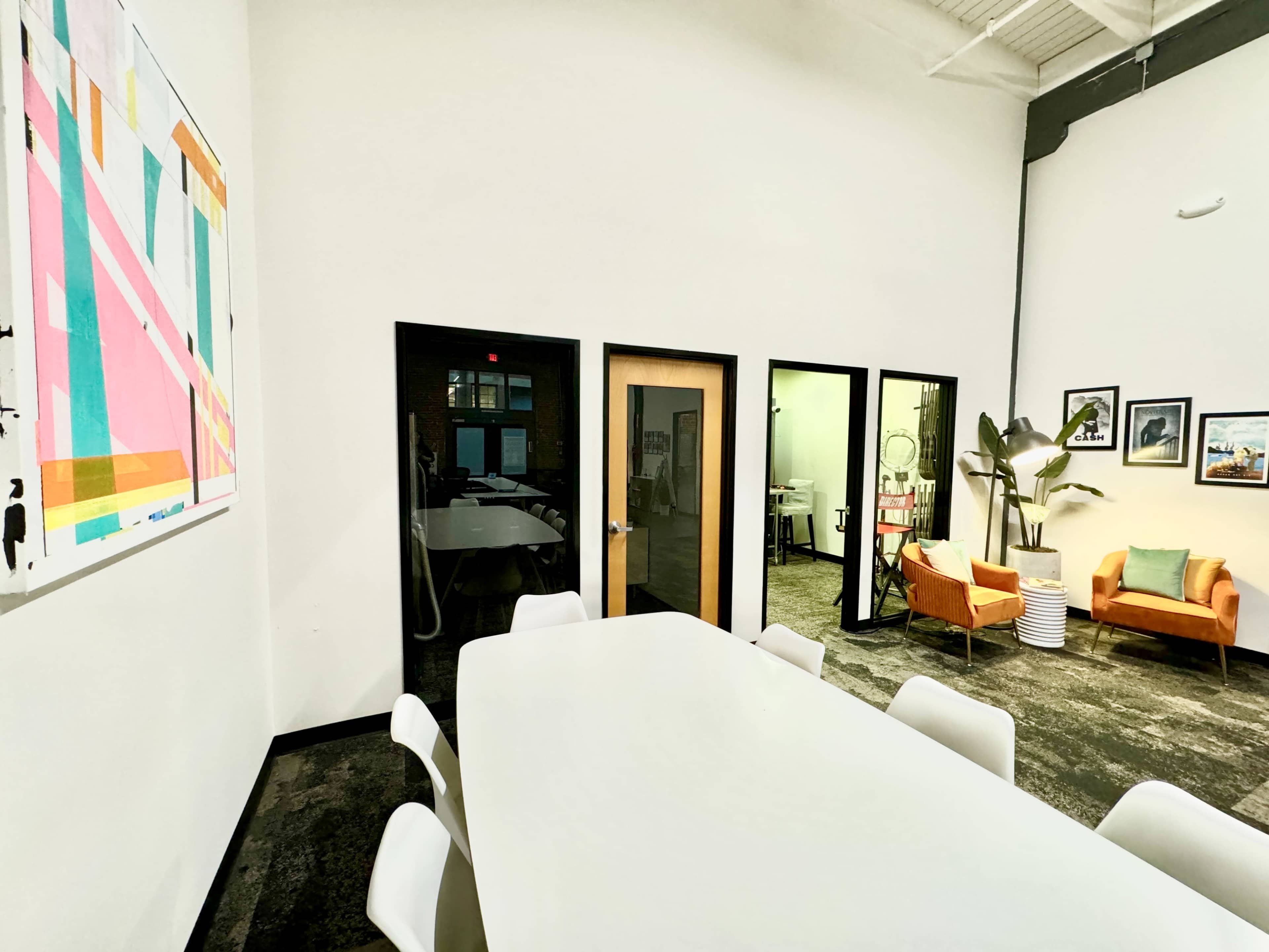 A bright, modern meeting room features a long white table, orange chairs, and several doors leading to different spaces, with artwork hanging on the wall.