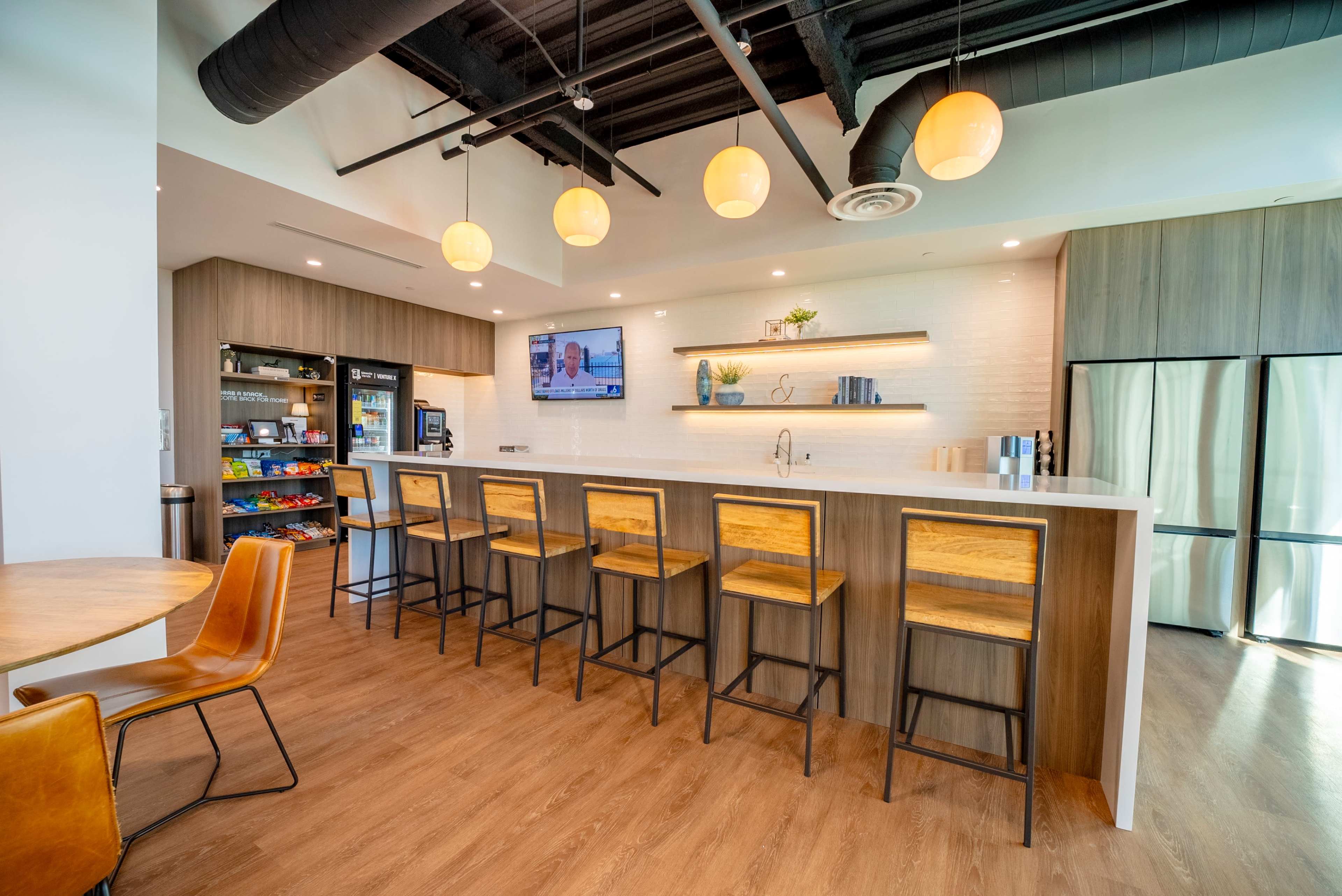 A modern kitchen area features a white bar counter with wooden stools, shelves displaying decorative items, and a large screen on the wall.