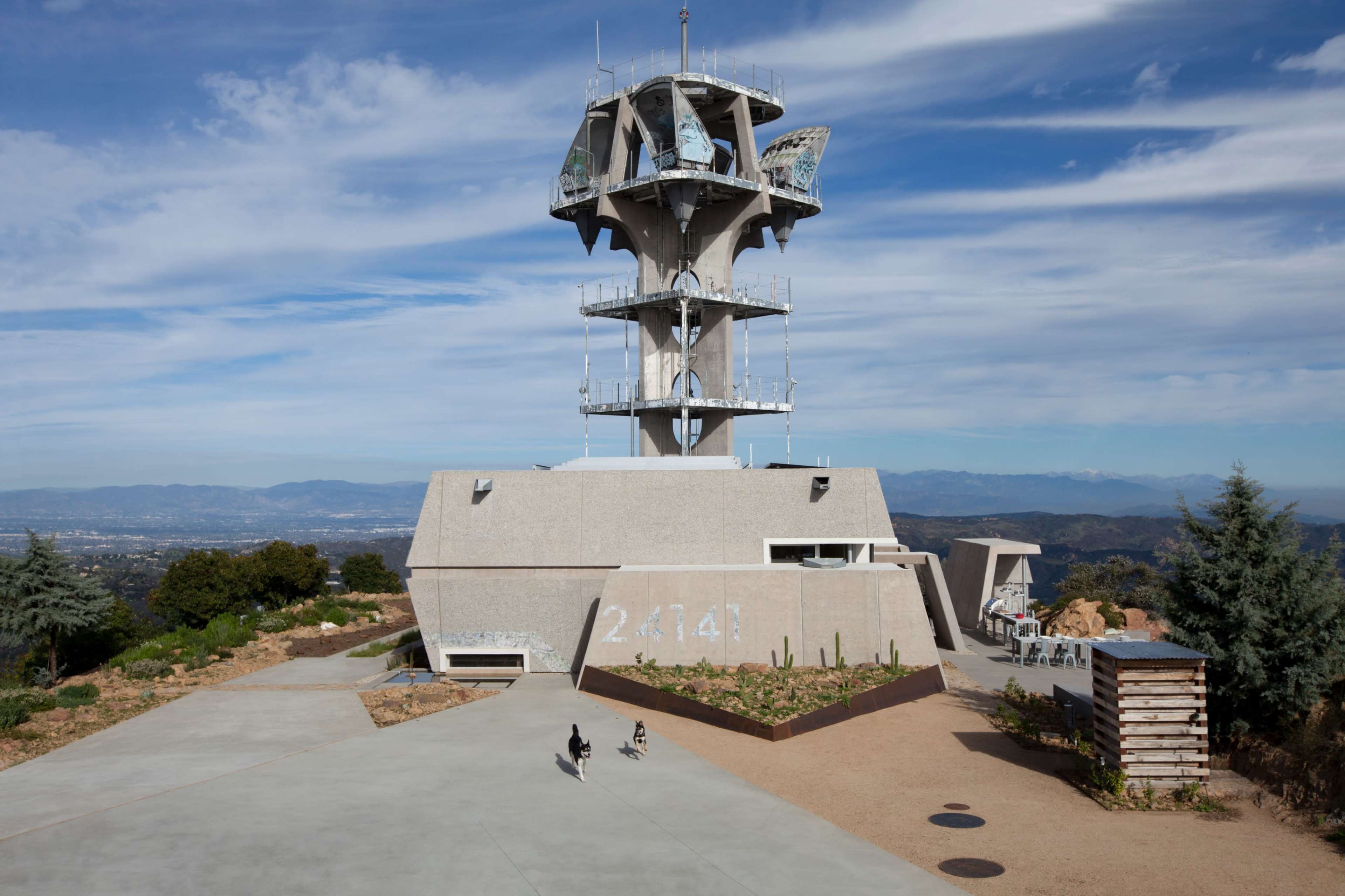 The image shows a modern tower structure with a helipad on top, surrounded by landscaped grounds and a clear sky.