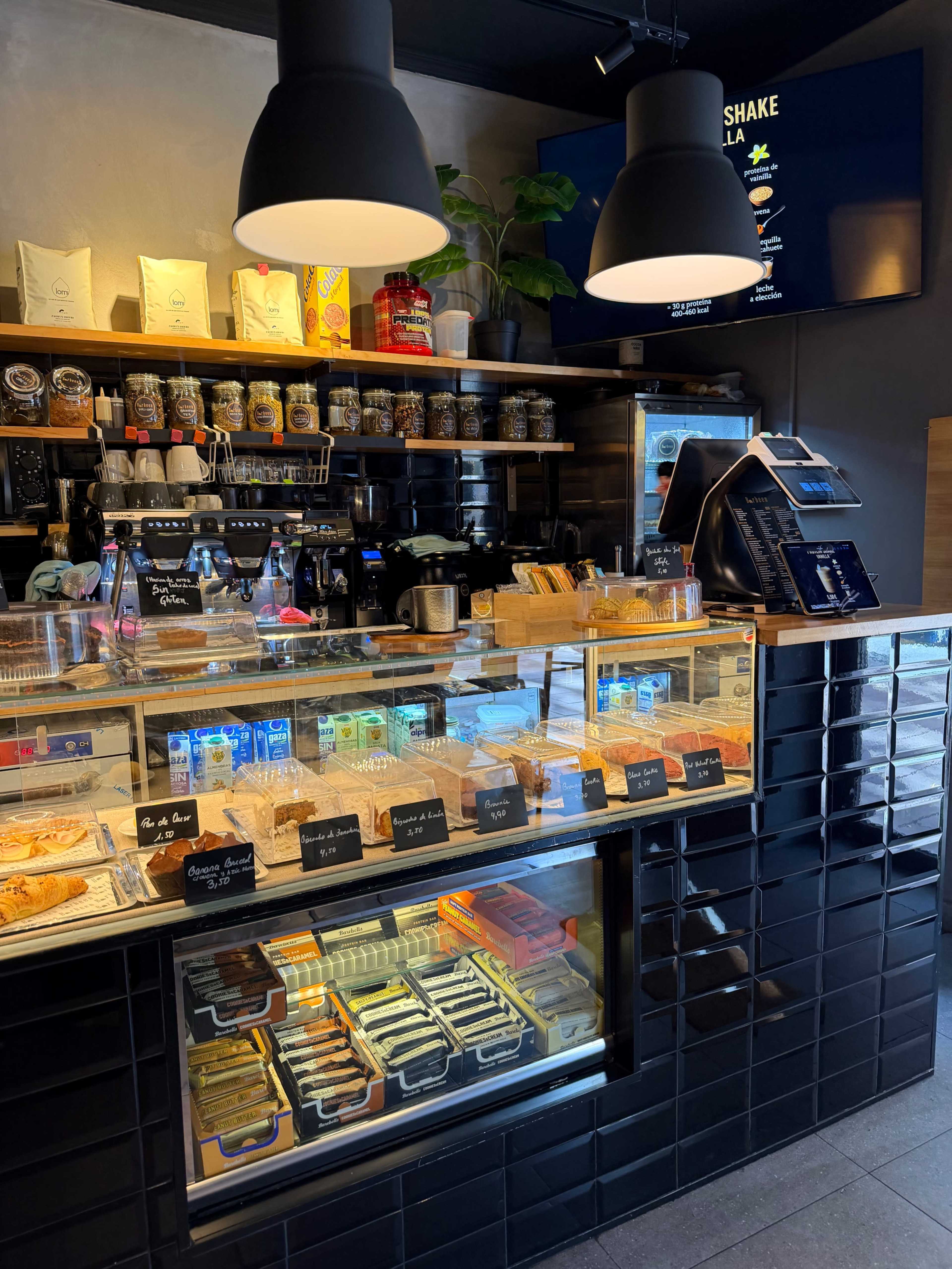The image shows a well-organized café counter featuring a selection of pastries, snacks, and beverages, along with various jars and equipment on shelves above.