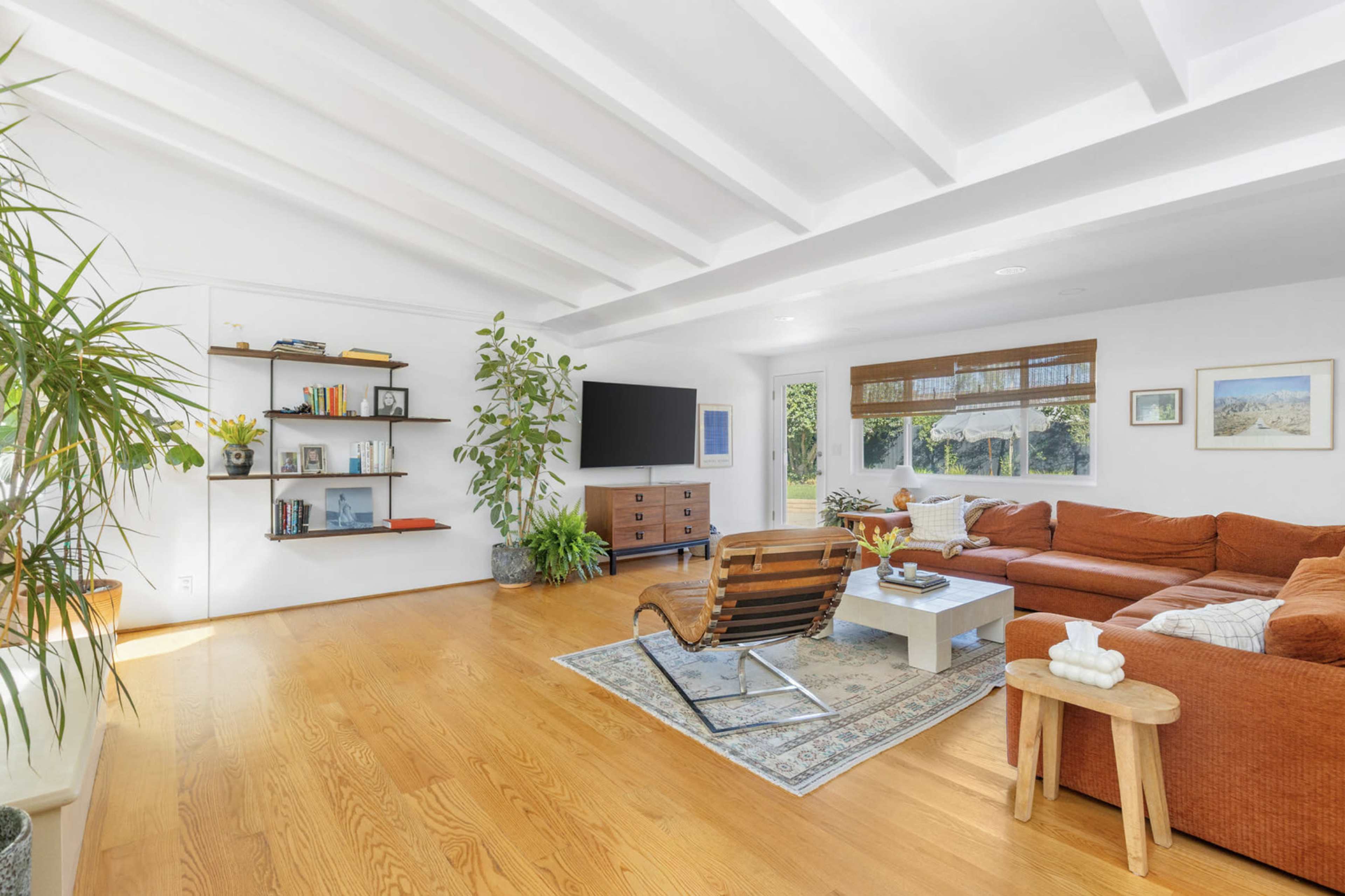 The living room features a large orange sectional sofa, a wooden coffee table, and a mounted television on a white wall, with plants and bookshelves adding decoration.